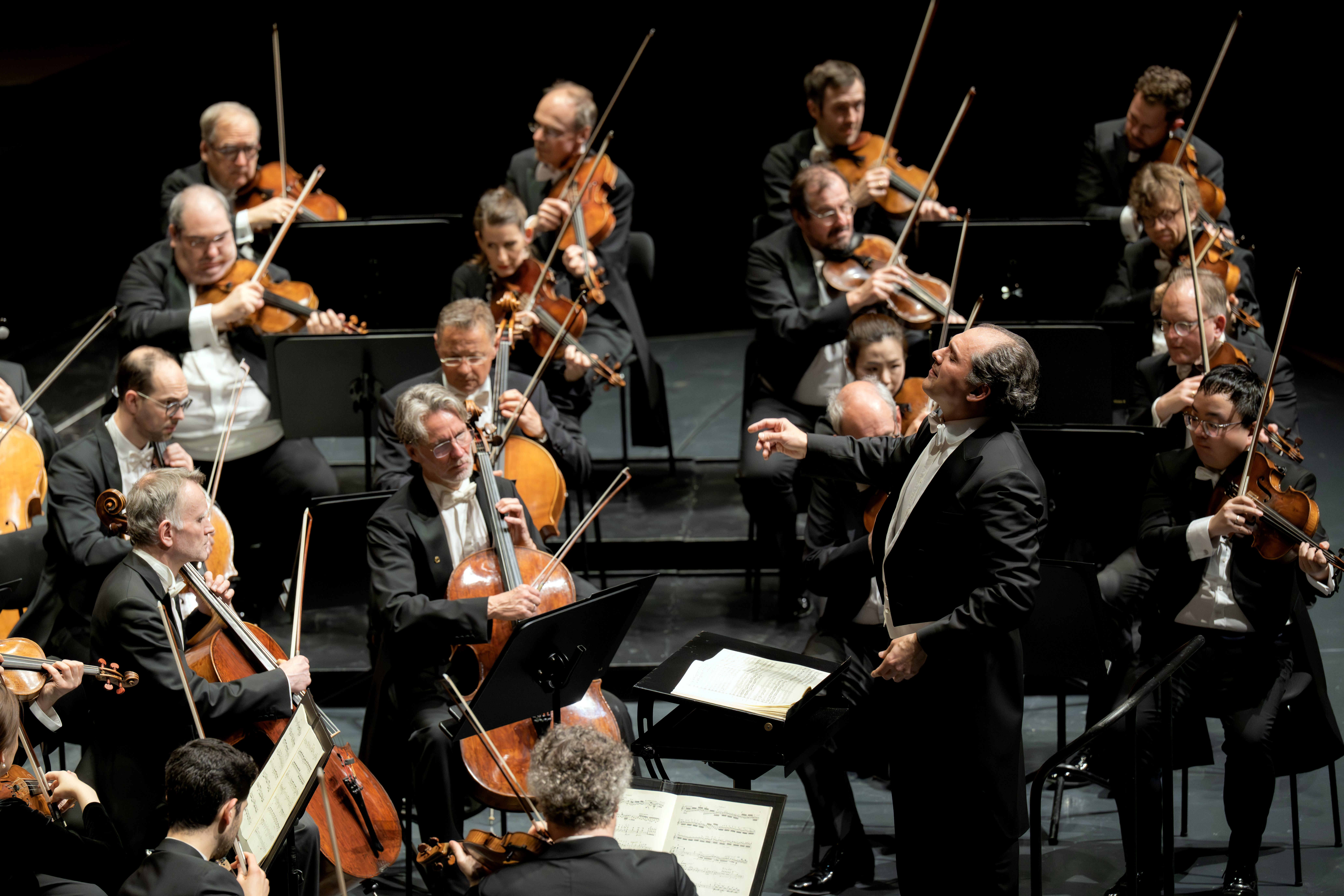 Ein Dirigent leitet ein Orchester von Musikern, die auf der Bühne Streichinstrumente spielen. Die Musiker, die förmlich in schwarze Anzüge gekleidet sind, konzentrieren sich im Bühnenlicht auf ihre Notenblätter und Instrumente.