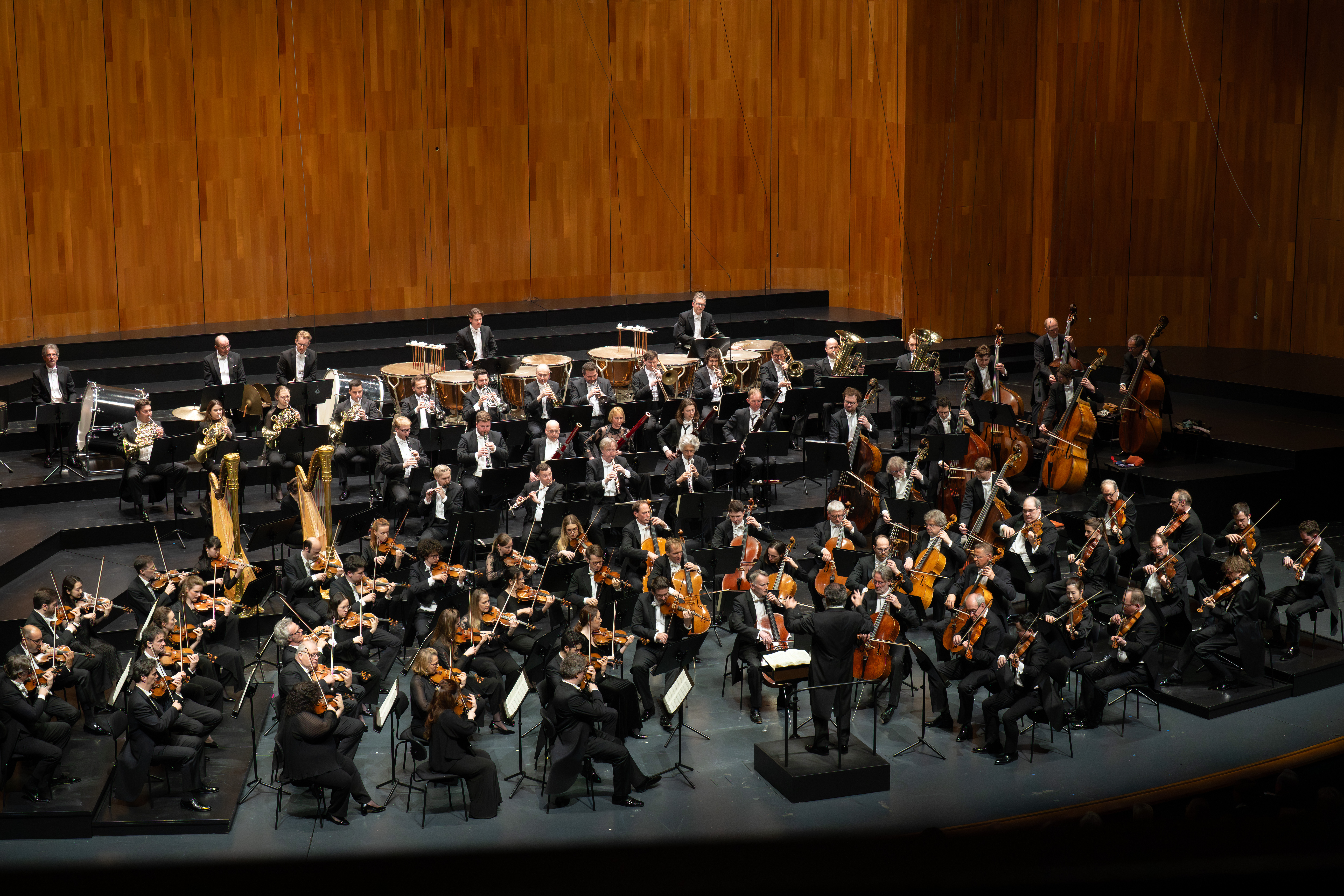 Auf der Bühne spielt ein großes Orchester mit Musikern, die Streicher, Holzbläser, Blechbläser und Schlaginstrumente spielen. Der Hintergrund ist holzgetäfelt, und der Dirigent steht auf dem mittleren Podium.