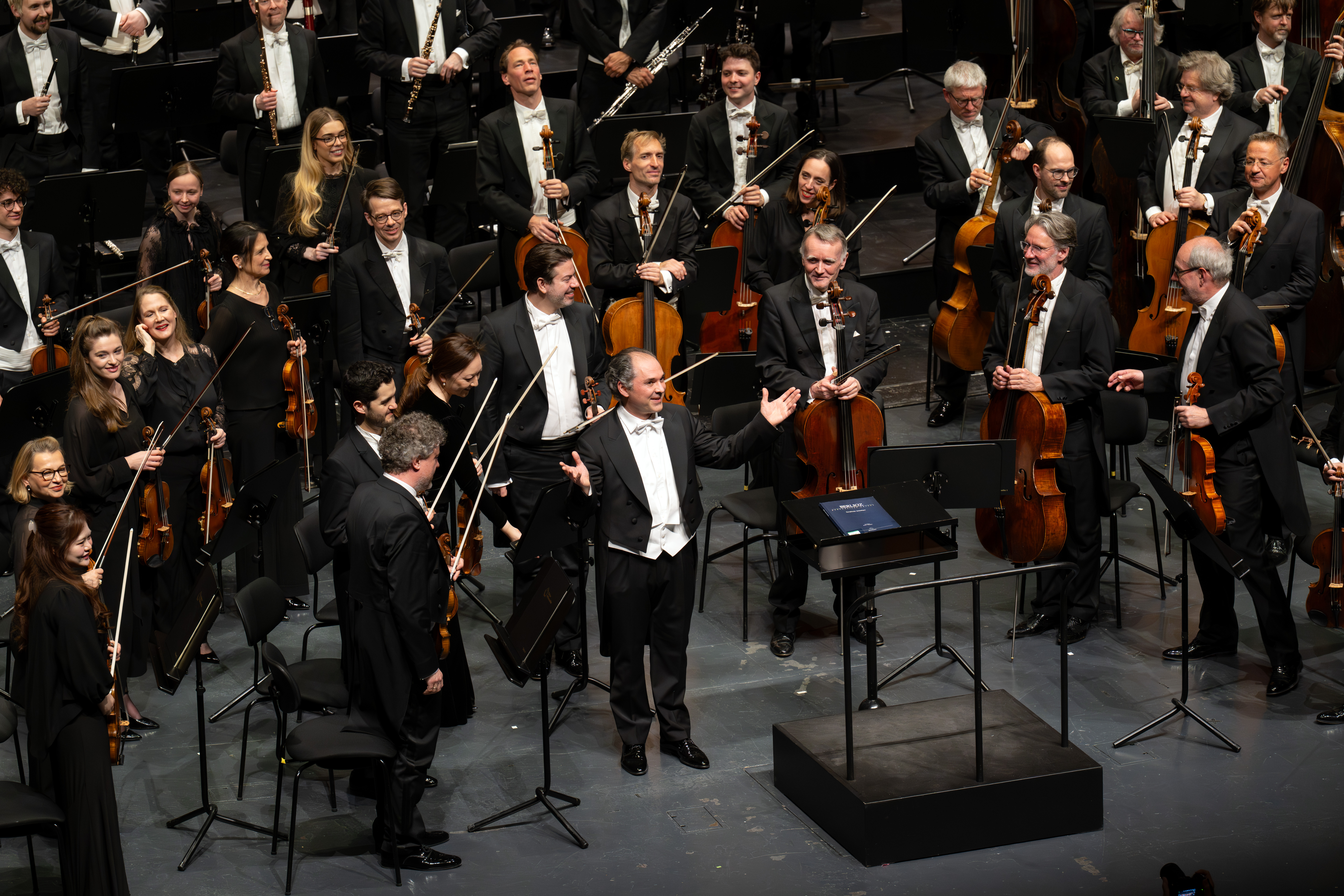 Tugan Sokhiev steht mit ausgestreckten Armen am Podium, während lächelnde Orchestermusiker mit Streichinstrumenten nach einer Aufführung auf der Bühne applaudieren und zu ihm hinschauen.