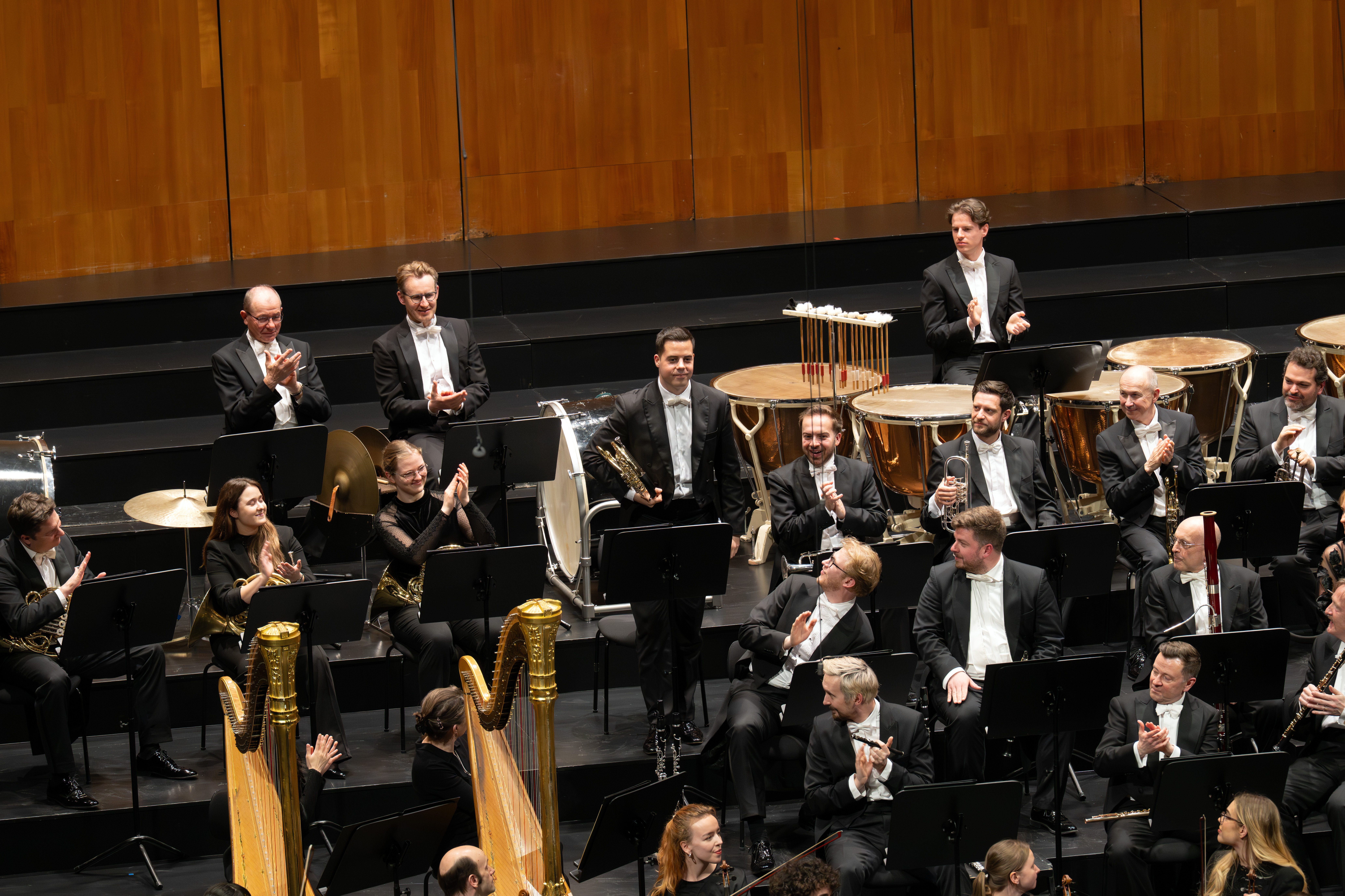 Ein Sinfonieorchester auf der Bühne, viele Musiker in schwarzen Smokings mit Instrumenten, einige stehen oder klatschen. Harfen, Blechbläser und Schlagzeug sind zu sehen. Der Hintergrund ist holzgetäfelt.