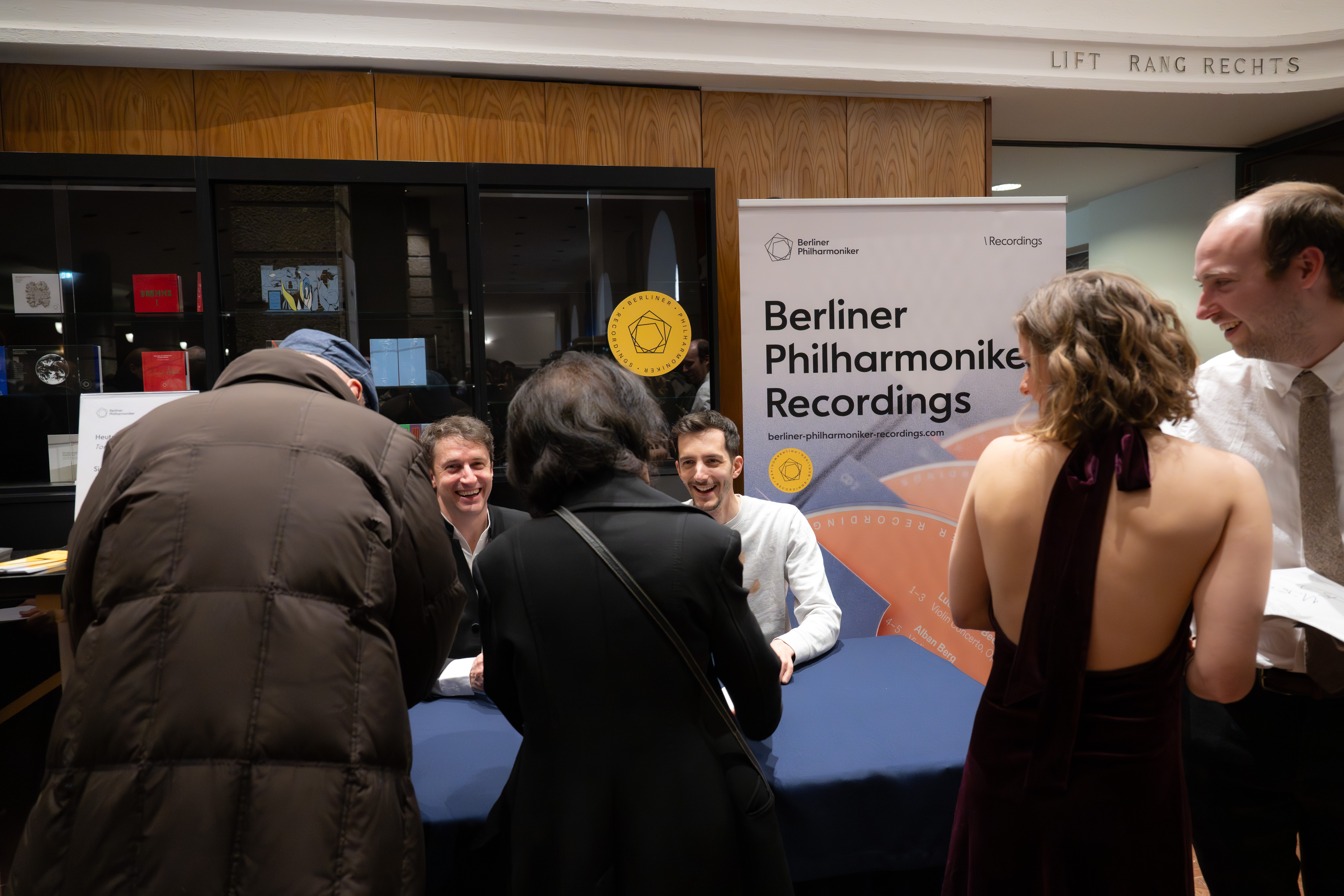 Zwei Männer sitzen an einem Tisch und schreiben Autogramme für Fans bei einer Veranstaltung der Berliner Philharmoniker Recordings. Eine Frau in einem burgunderroten Kleid und andere stehen am anderen Ende des Tisches, lächeln und unterhalten sich.