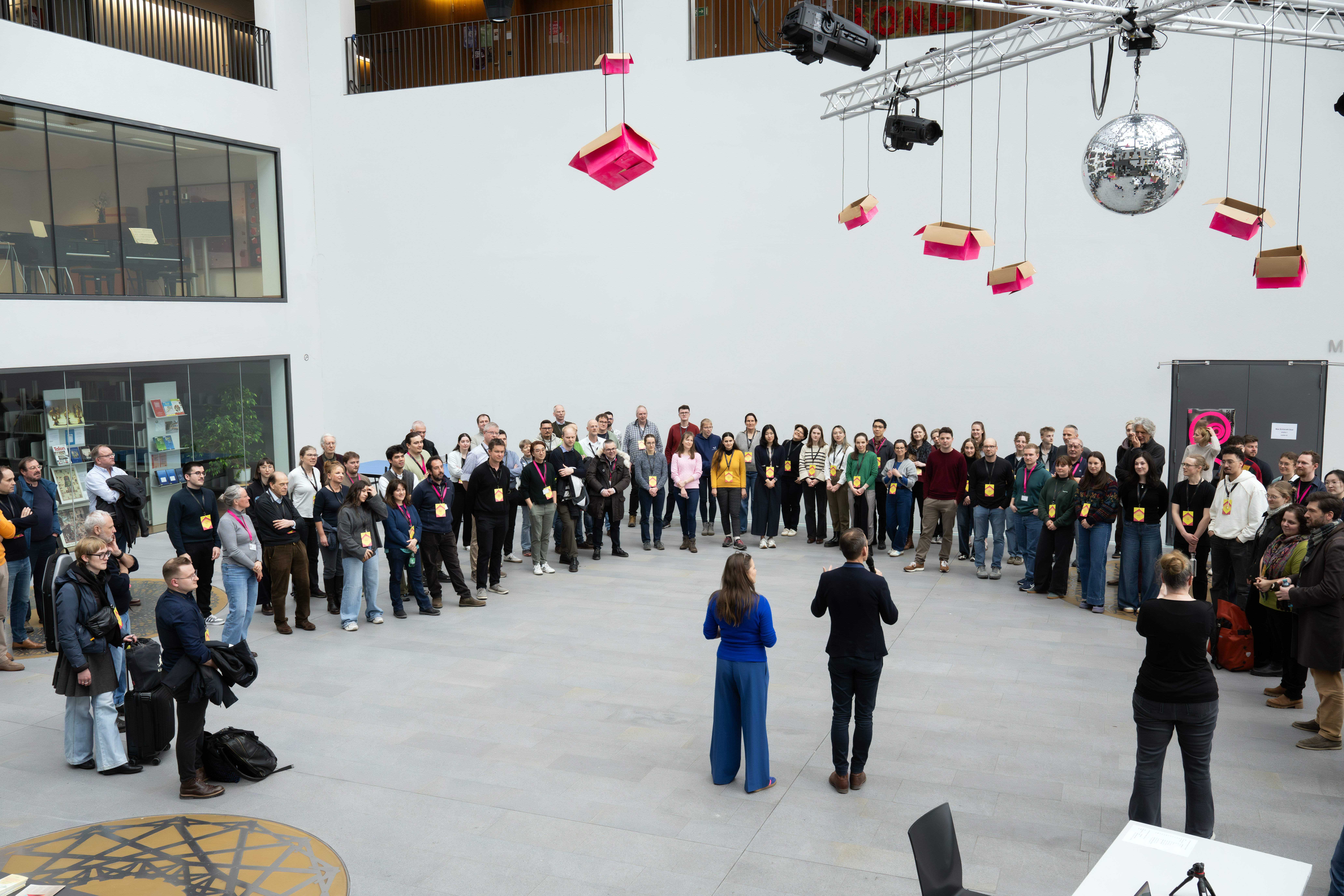 Eine große Gruppe von Menschen steht in einem Kreis in einem modernen, hellen Atrium und hört zwei Moderatoren in der Mitte zu. Von der Decke hängen rosa Boxen und eine Discokugel.