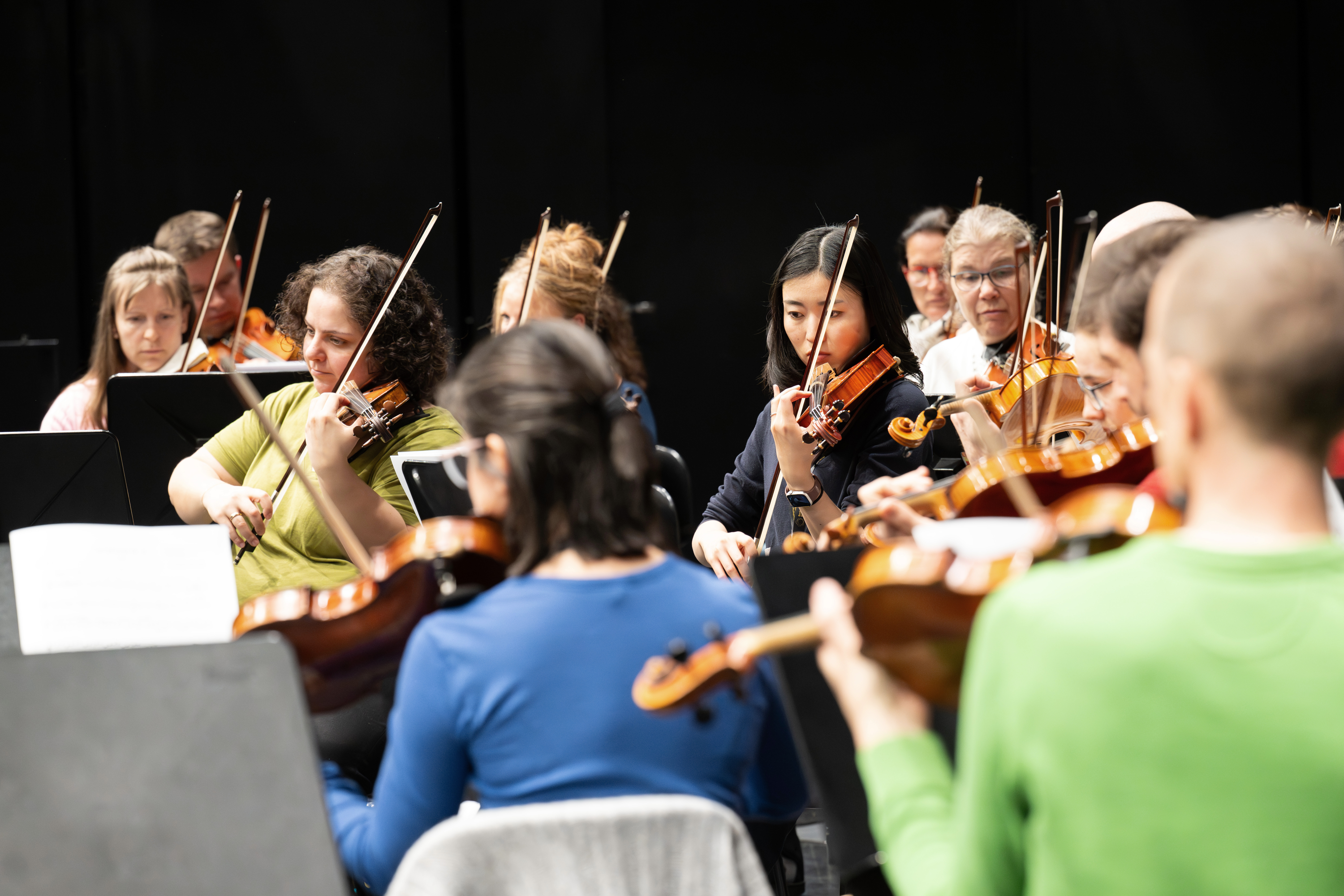 Eine Gruppe von Musikern spielt Geigen und Bratschen in einer Orchesterprobe, konzentriert auf ihre Notenblätter, wobei ein dunkler Hintergrund ihre Konzentration und ihre Instrumente hervorhebt.