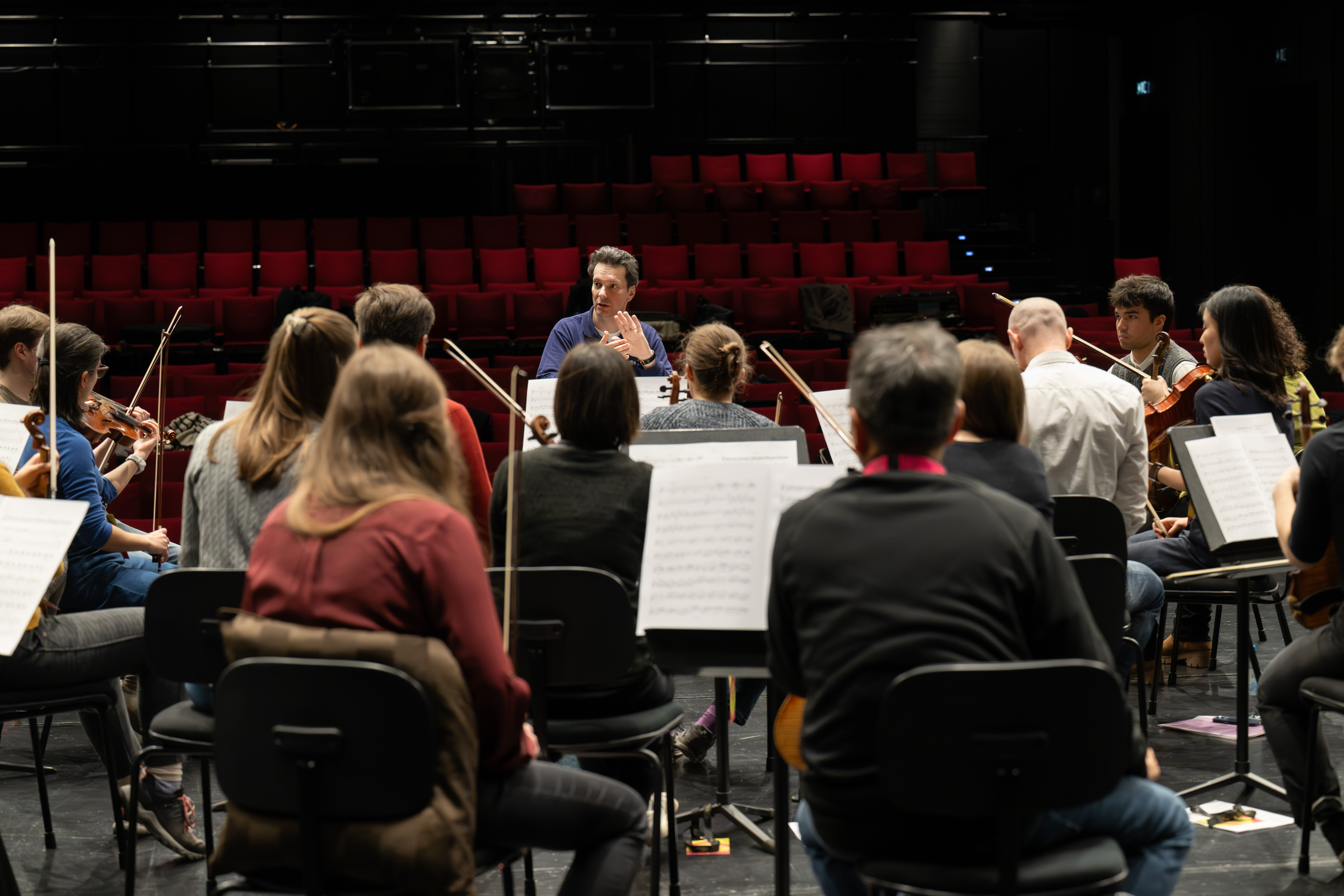 Ein Dirigent leitet eine Probe mit einem Orchester, das in einem Halbkreis auf einer Bühne sitzt. Notenblätter sind zu sehen, und im Hintergrund sind leere rote Theatersitze zu sehen.