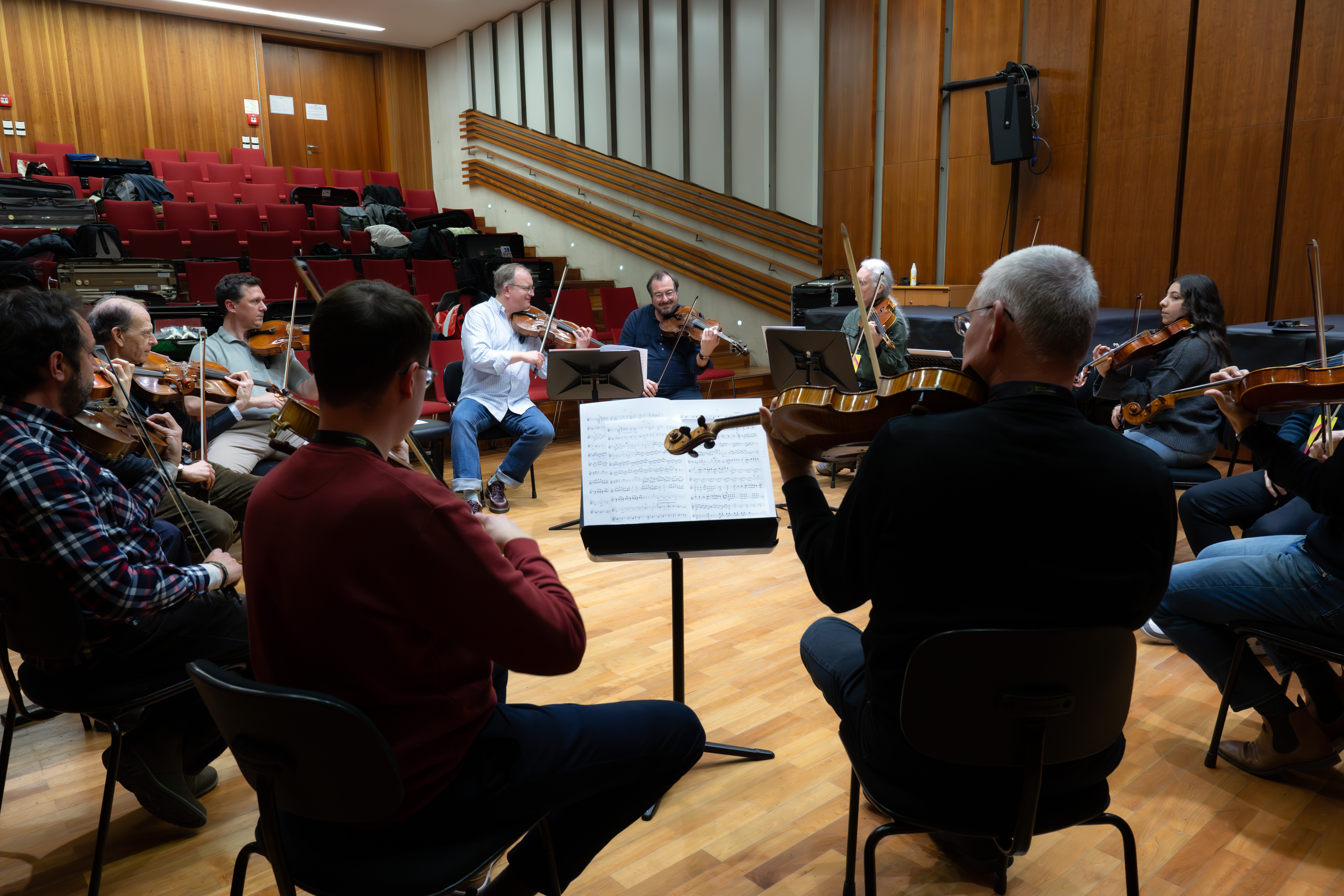 Eine Gruppe von Musikern sitzt im Halbkreis in einem Proberaum, spielt auf Streichinstrumenten und liest Noten, im Hintergrund leere rote Stühle und Holzwände.