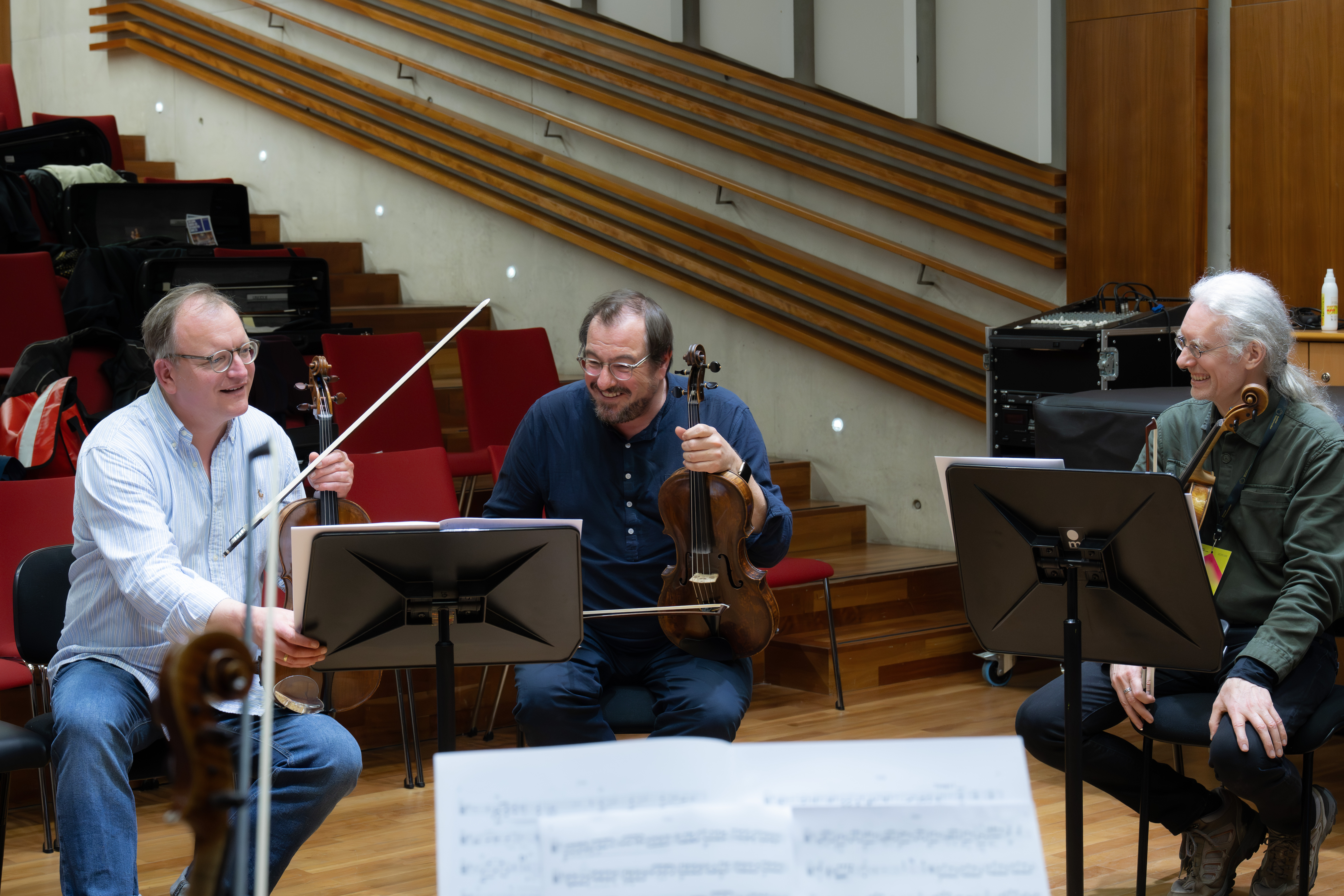 Drei Musiker mit Streichinstrumenten sitzen lächelnd in einem Proberaum mit roten Stühlen, Holzböden und Notenständern zusammen und scheinen einen unbeschwerten Moment zu genießen.