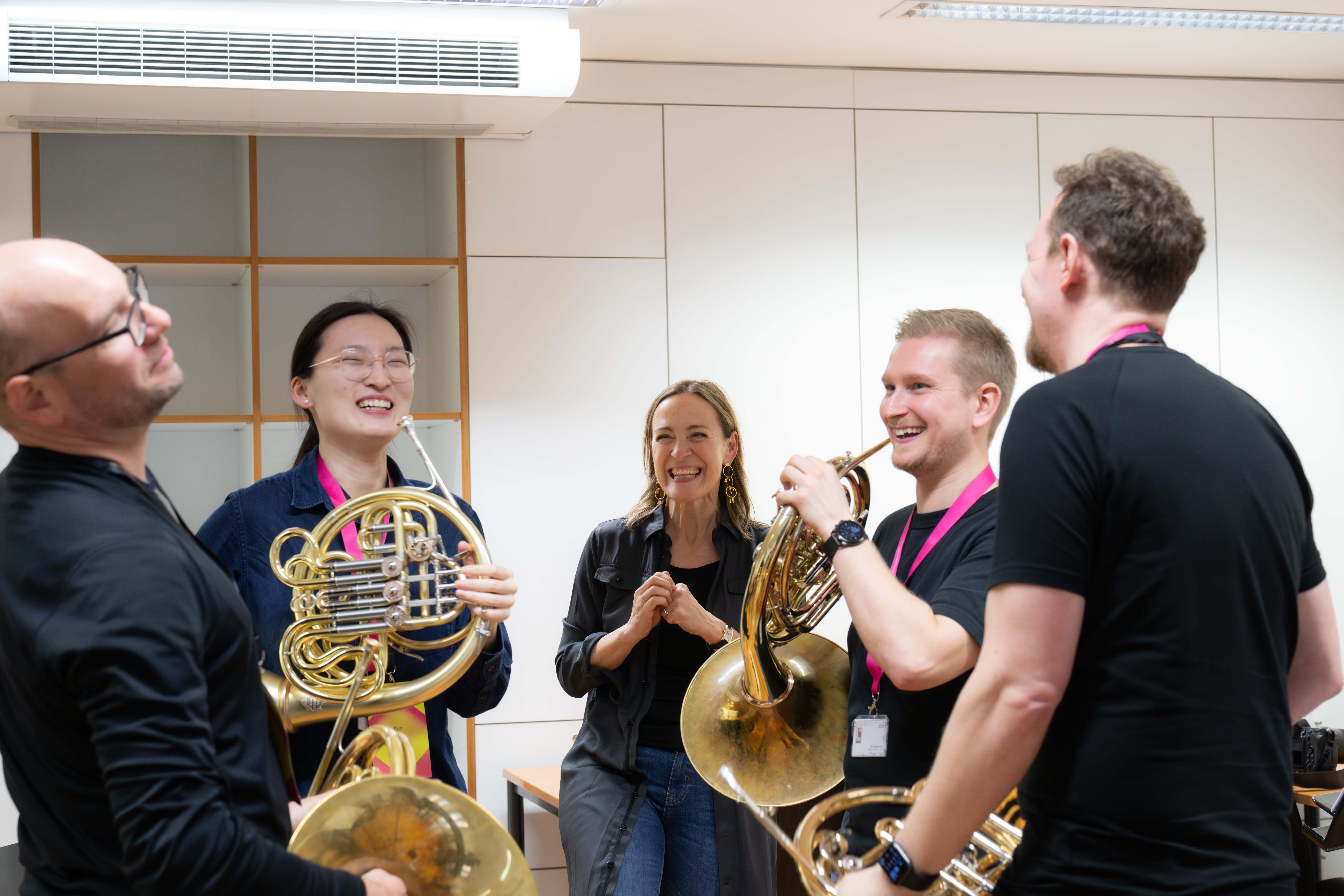Vier Personen, die Waldhörner in der Hand halten, lachen gemeinsam in einem hellen Raum, während eine Frau im Hintergrund lächelt und sie beobachtet. Sie alle scheinen einen unbeschwerten Moment während einer Probe zu genießen.