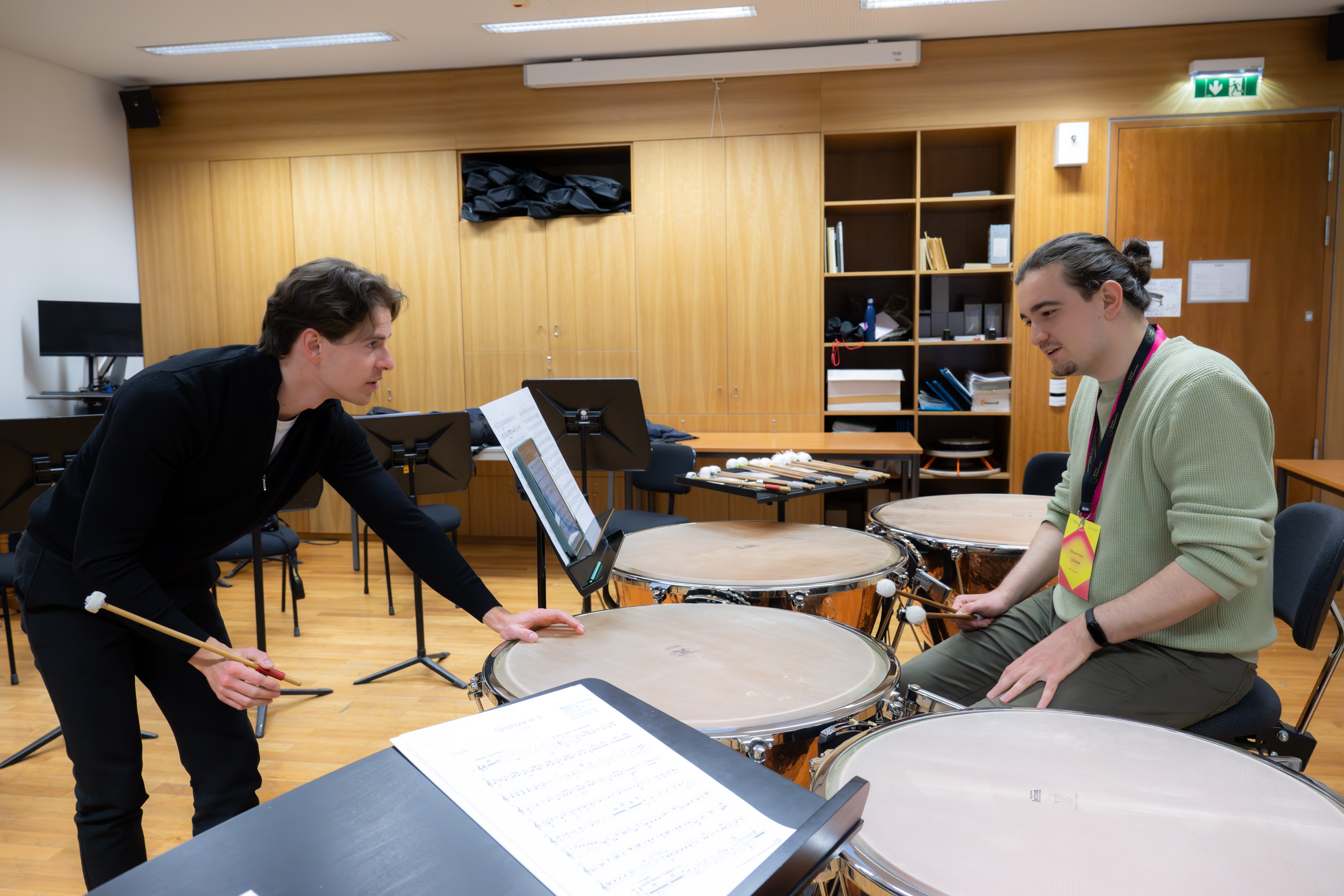 Zwei Personen befinden sich in einem Musikunterrichtsraum mit Pauken und Trommeln. Eine Person steht und spricht, während sie die Trommelstöcke hält; die andere sitzt und hört zu. Um sie herum sind Notenblätter und Notenständer zu sehen.