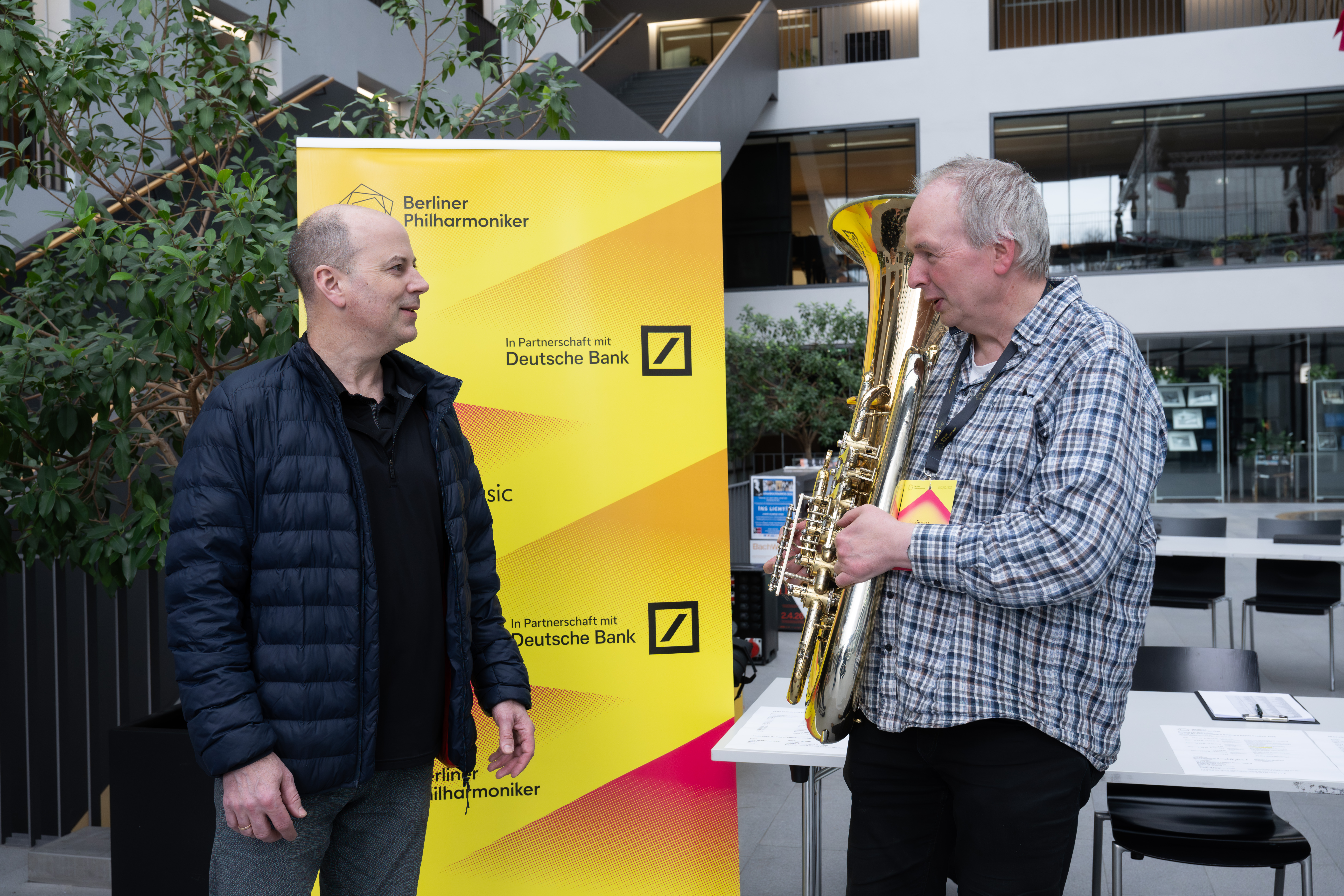 Zwei Männer stehen in einem Innenraum und unterhalten sich; einer hält ein Blechblasinstrument. Hinter ihnen hängt ein gelbes Banner der Berliner Philharmoniker mit den Logos der Deutschen Bank. Die Umgebung wirkt modern, mit Pflanzen und offenem Raum.