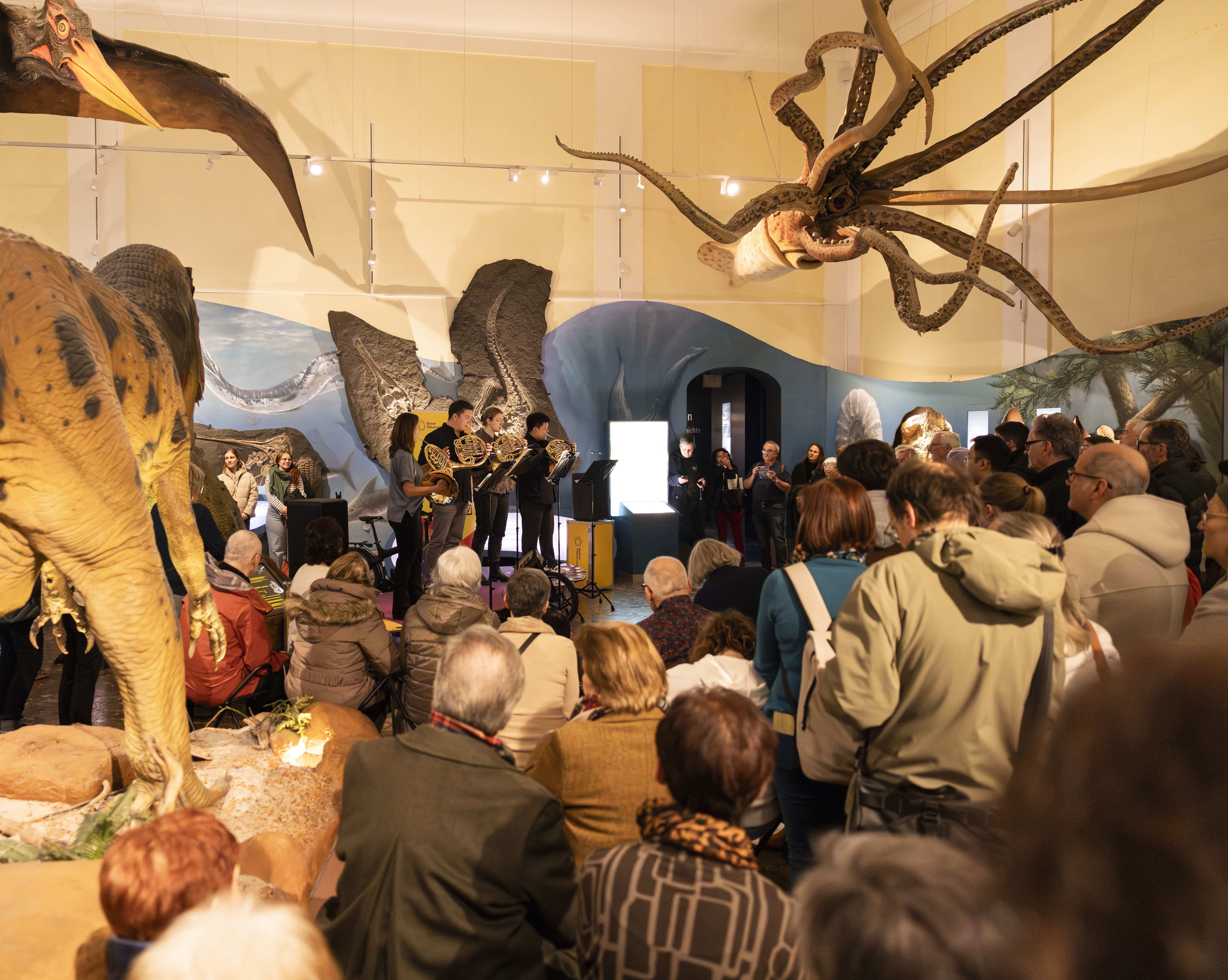 A crowd watches a small brass ensemble perform in a museum, surrounded by large prehistoric animal exhibits, including a dinosaur and a giant squid model hanging from the ceiling.