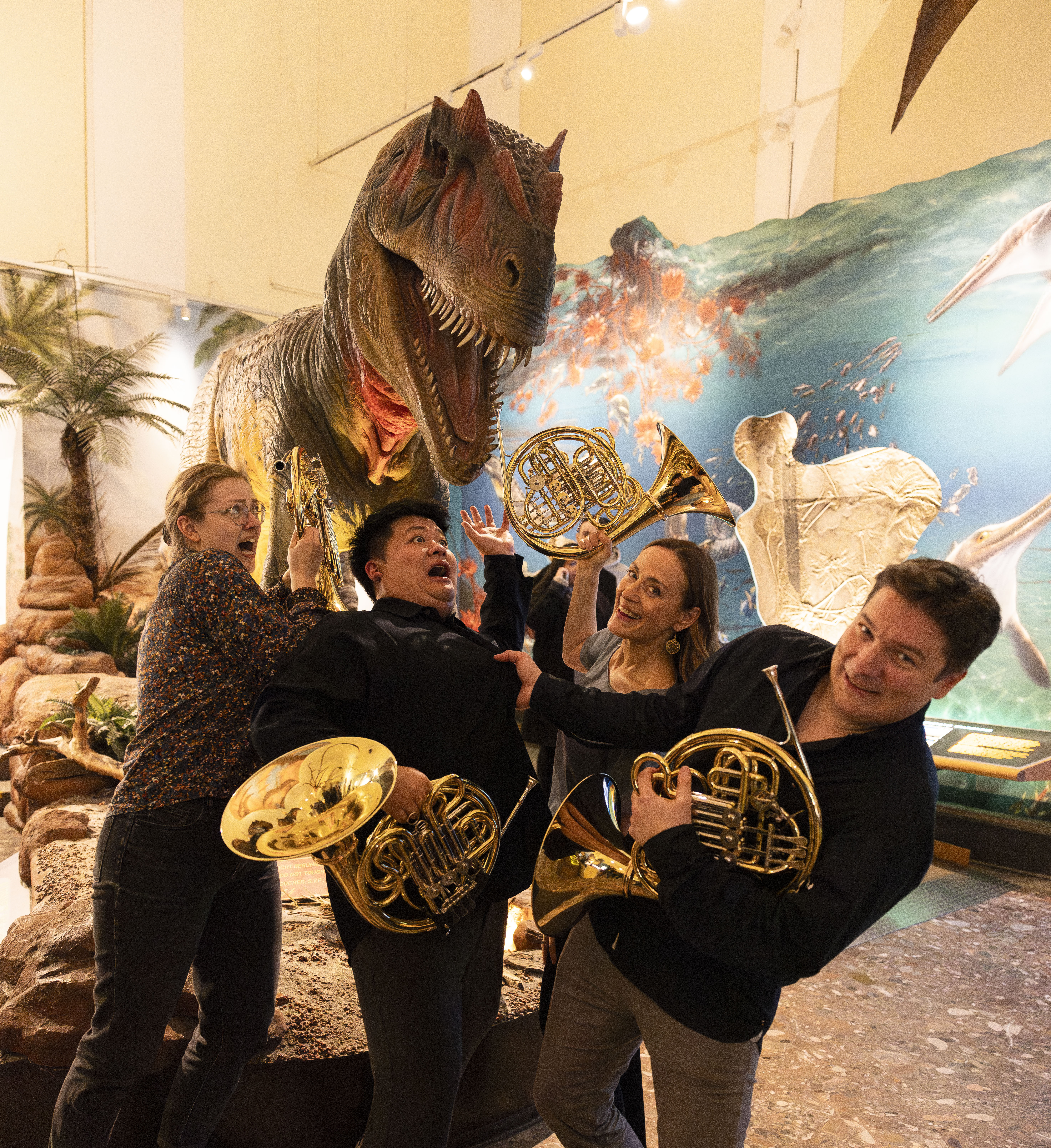 Four people holding French horns pose playfully in front of a large, open-mouthed dinosaur model in a museum, pretending to be startled or attacked by the dinosaur.