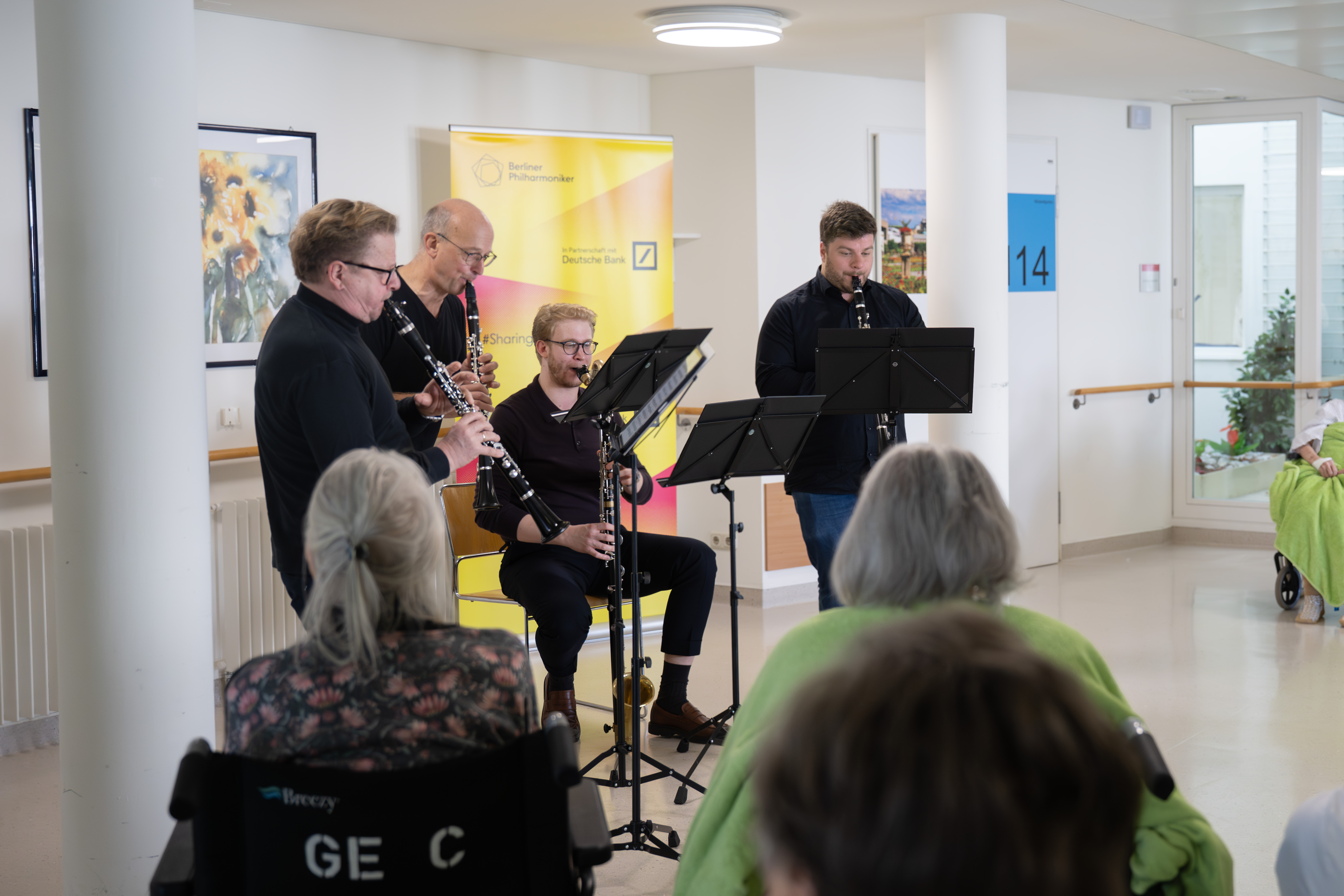 Four musicians play clarinets in front of an audience in a bright room, with some listeners seated in wheelchairs. A yellow banner and flower paintings are visible in the background.