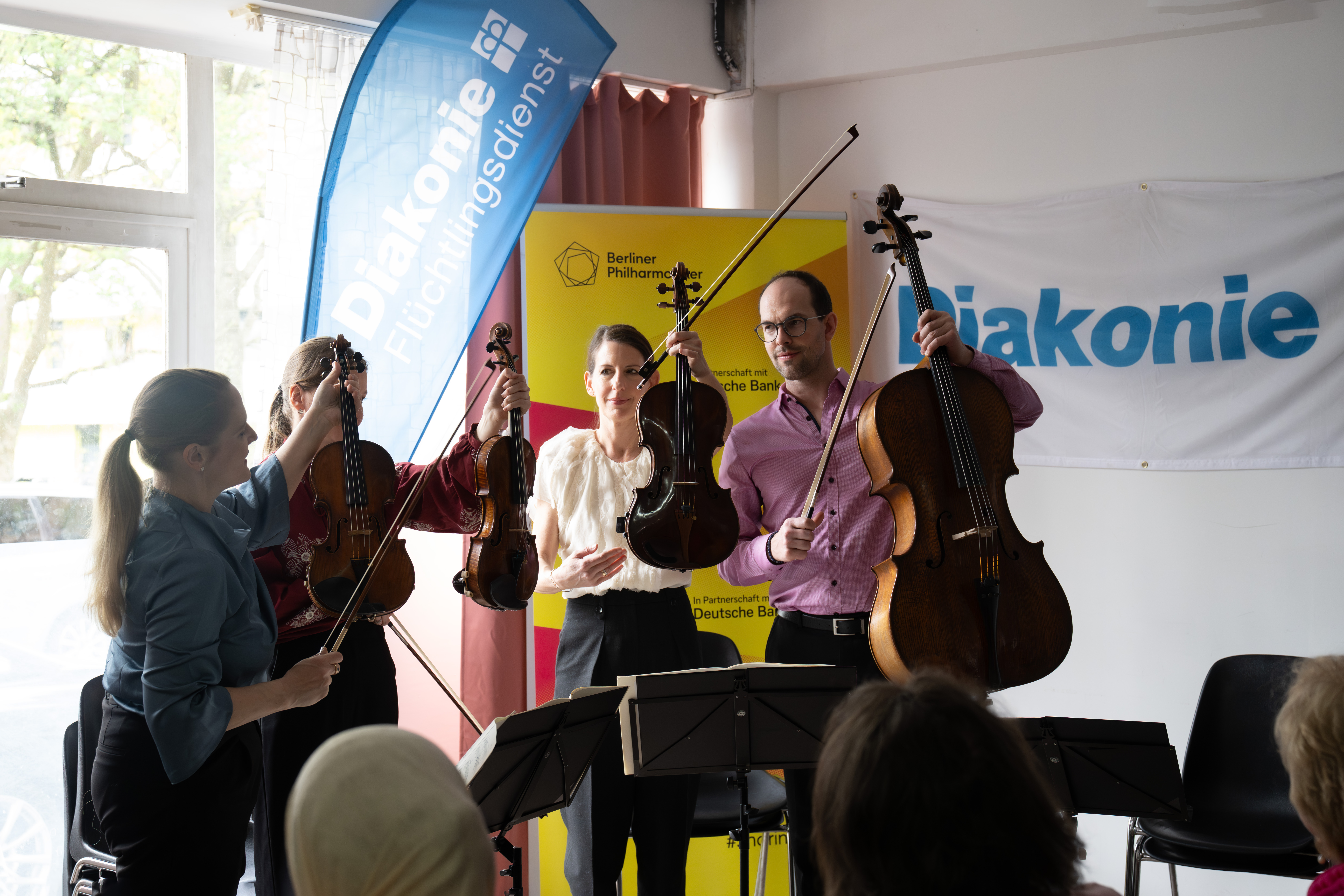 Four musicians holding string instruments stand in front of Diakonie and Berliner Philharmoniker banners, smiling and interacting, with sheet music stands and audience members visible in the foreground.