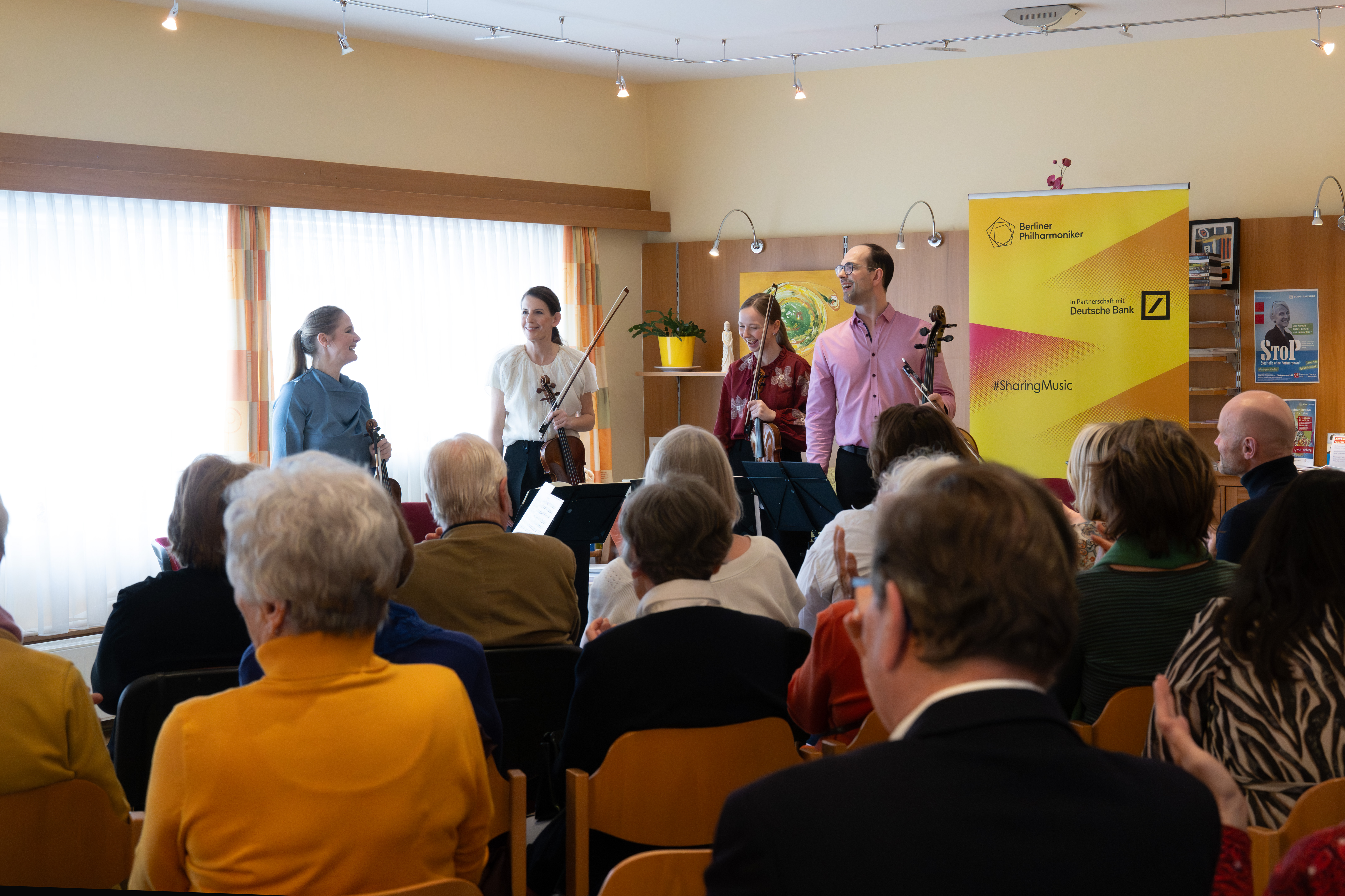 Four musicians stand at the front of a room, holding string instruments and smiling at an audience. People are seated, watching the performers. A yellow banner in the background reads “Sharing Music.”.