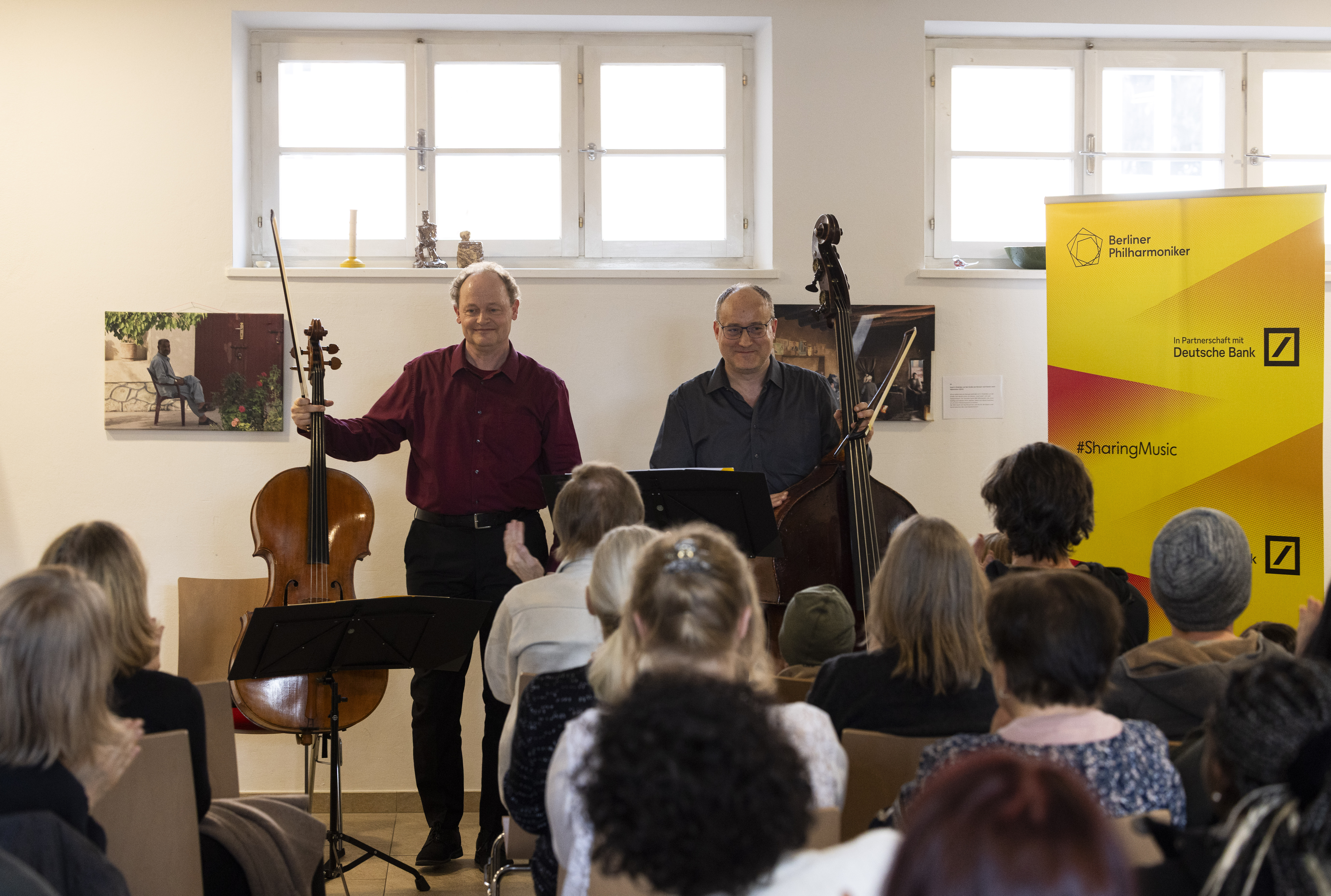 Two musicians stand with a cello and double bass, smiling at an applauding seated audience in a bright room. A yellow Berliner Philharmoniker banner is visible on the right.