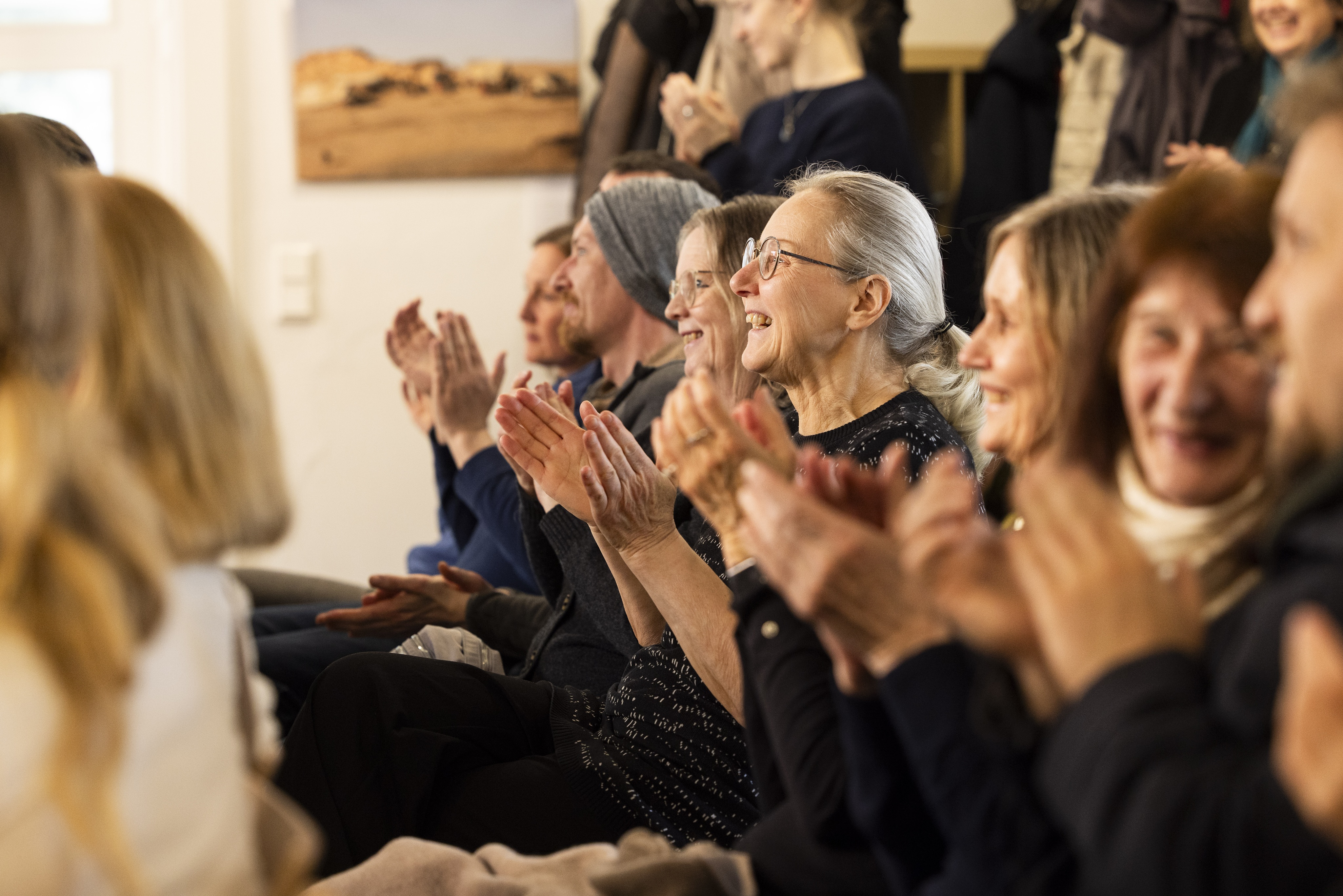 A group of people sit closely together indoors, smiling and enthusiastically clapping, suggesting they are enjoying a performance or presentation. The mood is warm and joyful.