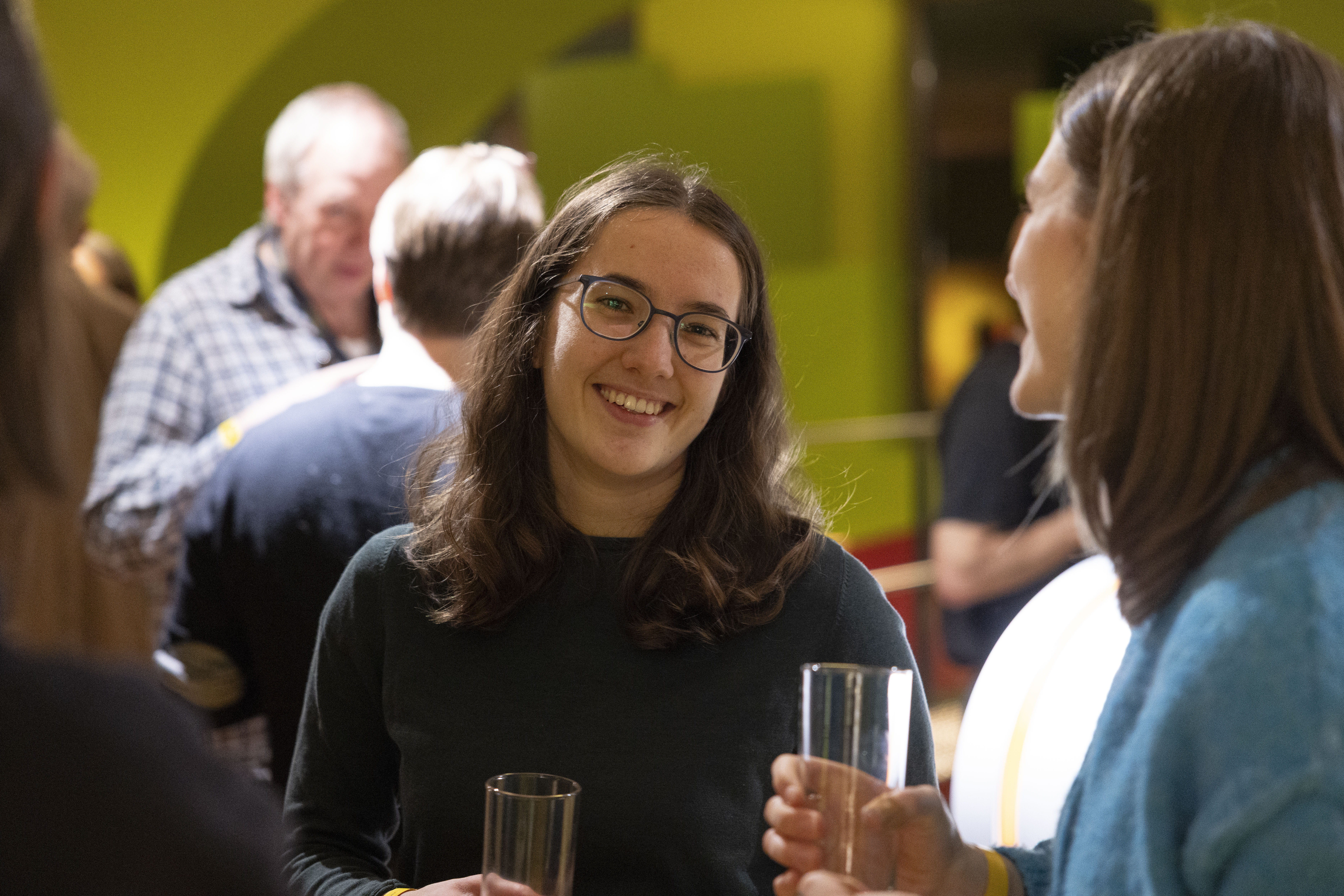 Eine Frau mit Brille lächelt in die Kamera und hält ein Glas bei einem geselligen Beisammensein. Im Hintergrund, bei warmer Beleuchtung und grünen Wänden, unterhalten sich Menschen und mischen sich.