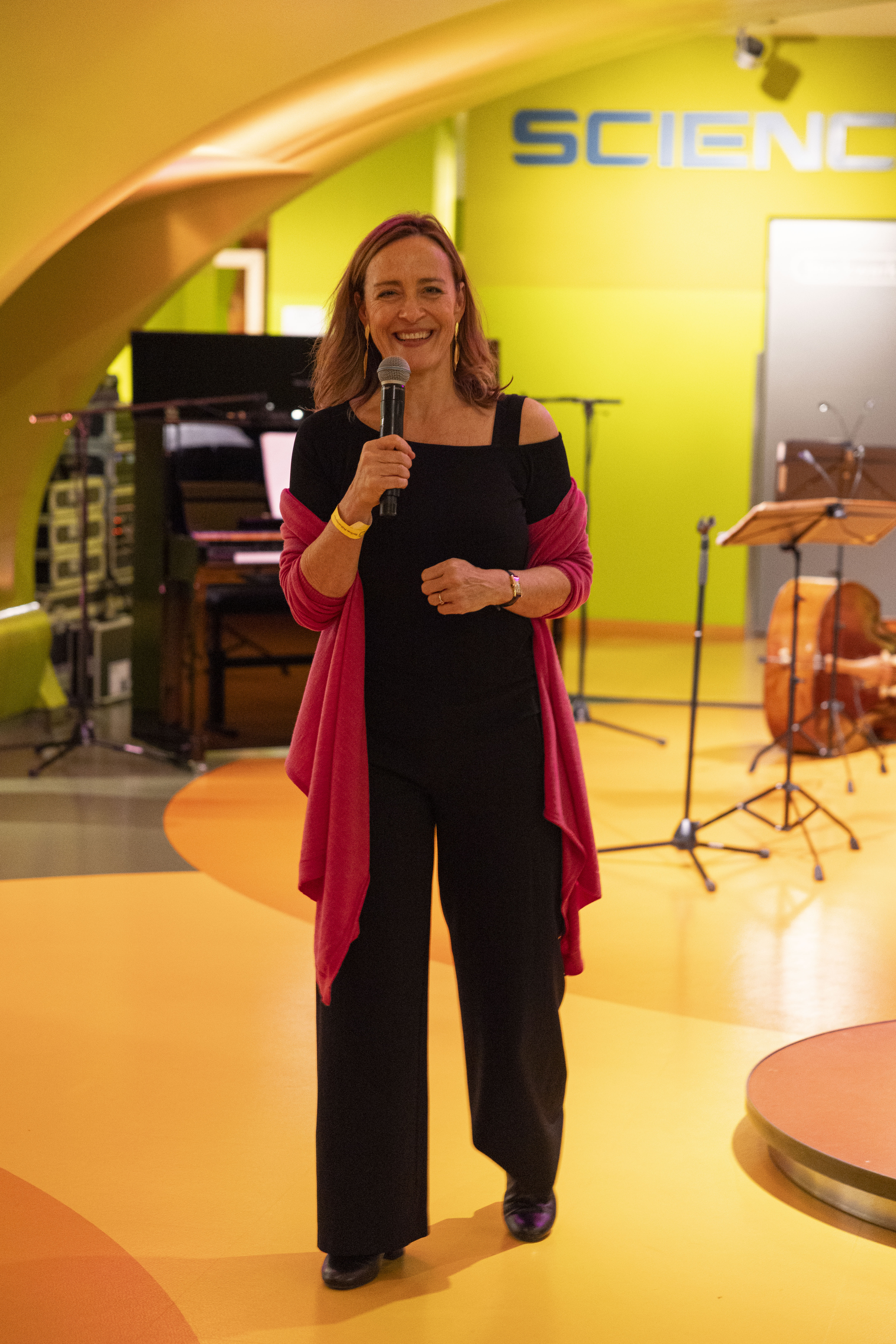 Sarah Willis in schwarzer Kleidung und einem rosa Schal hält ein Mikrofon und lächelt, während sie auf einer bunten Bühne mit Musikinstrumenten und einem Schild mit der Aufschrift SCIENCE im Hintergrund steht.