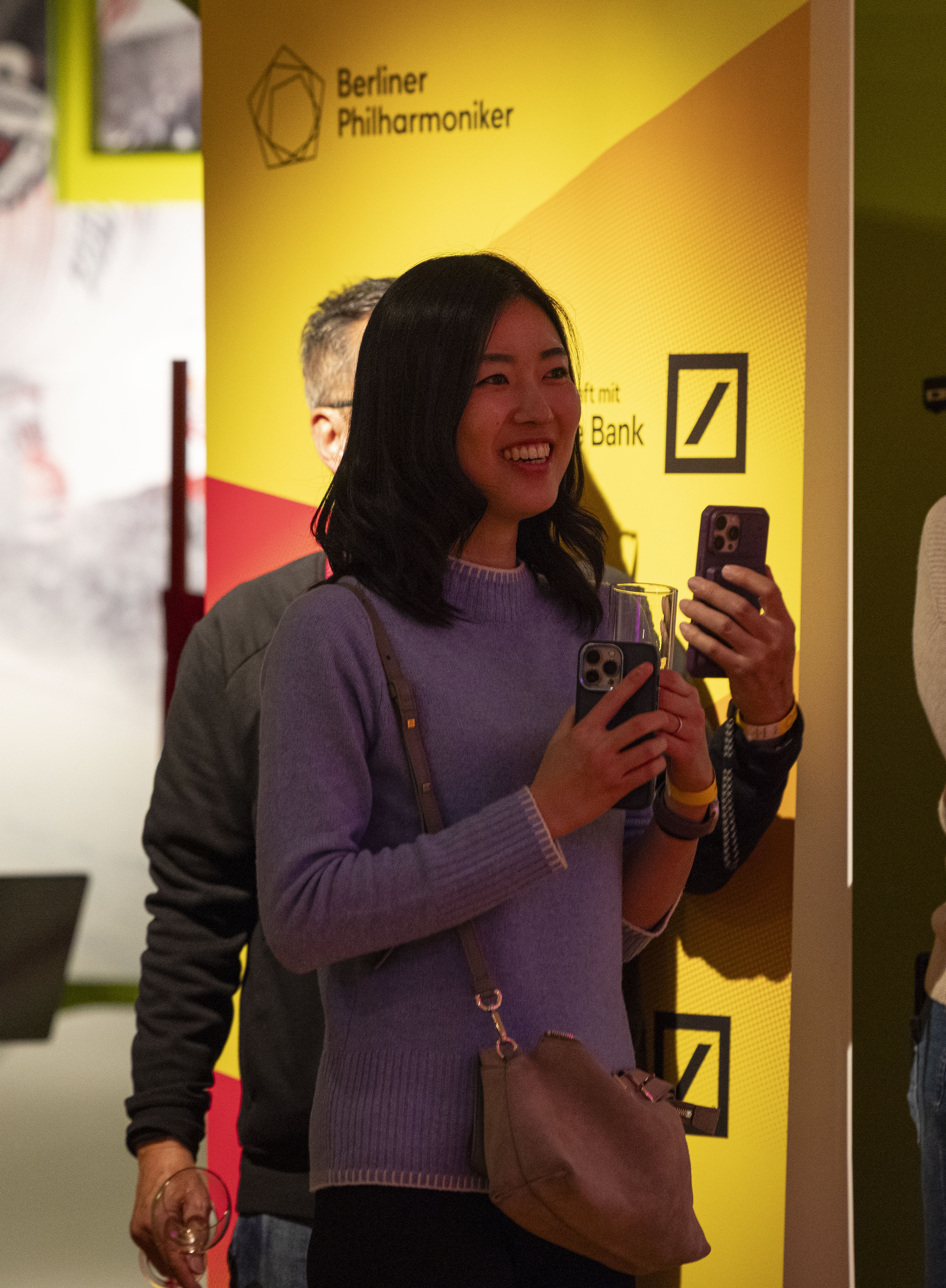 Eine Frau in einem lila Pullover lächelt, während sie ein Getränk und ein Telefon in der Hand hält. Sie steht neben einer gelben Wand mit den Logos der Berliner Philharmoniker und der Deutschen Bank im Hintergrund.