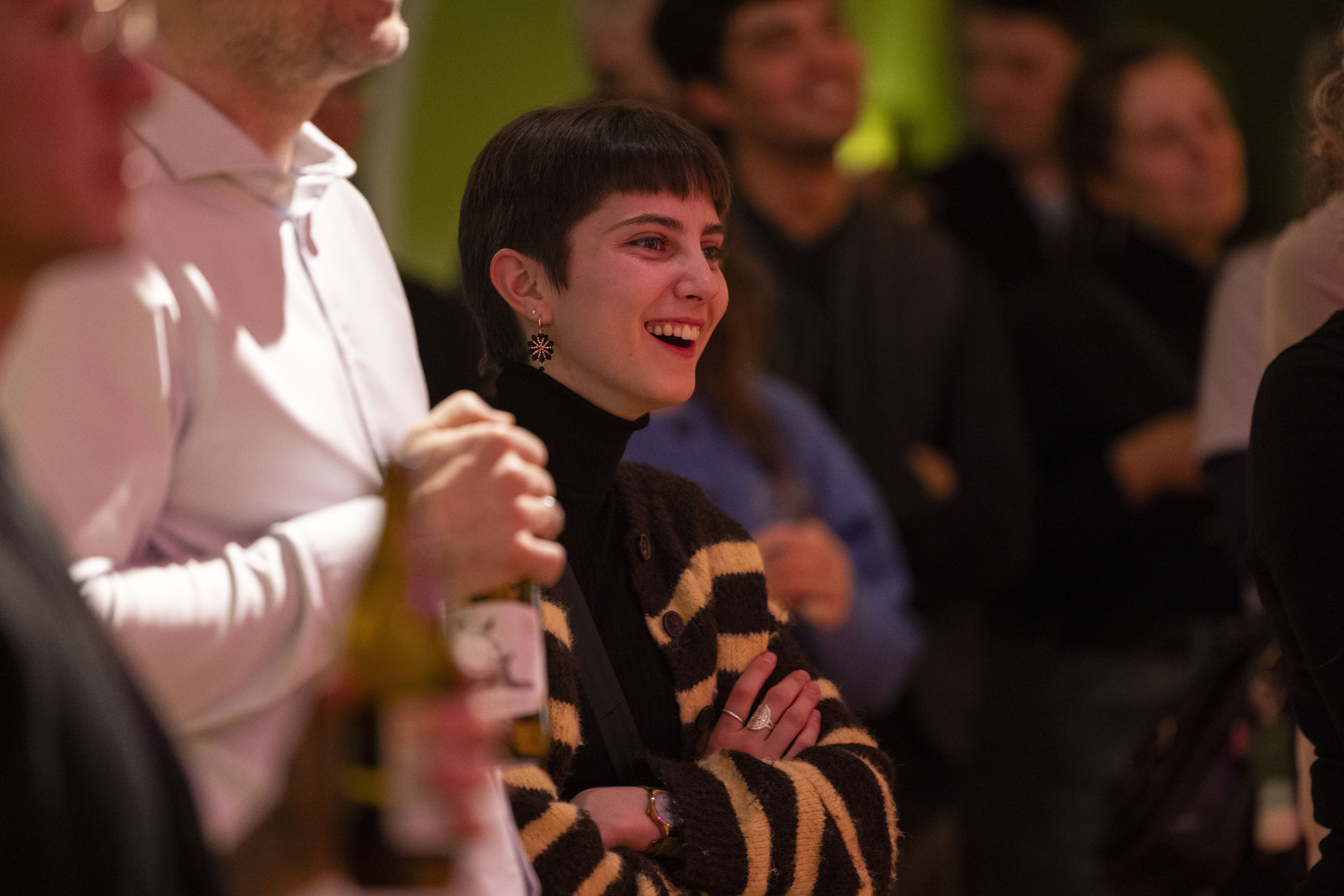 Eine Person mit kurzen dunklen Haaren und einem gestreiften Pullover lächelt mit verschränkten Armen in einer Menschenmenge, wirkt engagiert und glücklich; andere Personen um sie herum sind im Hintergrund verschwommen.