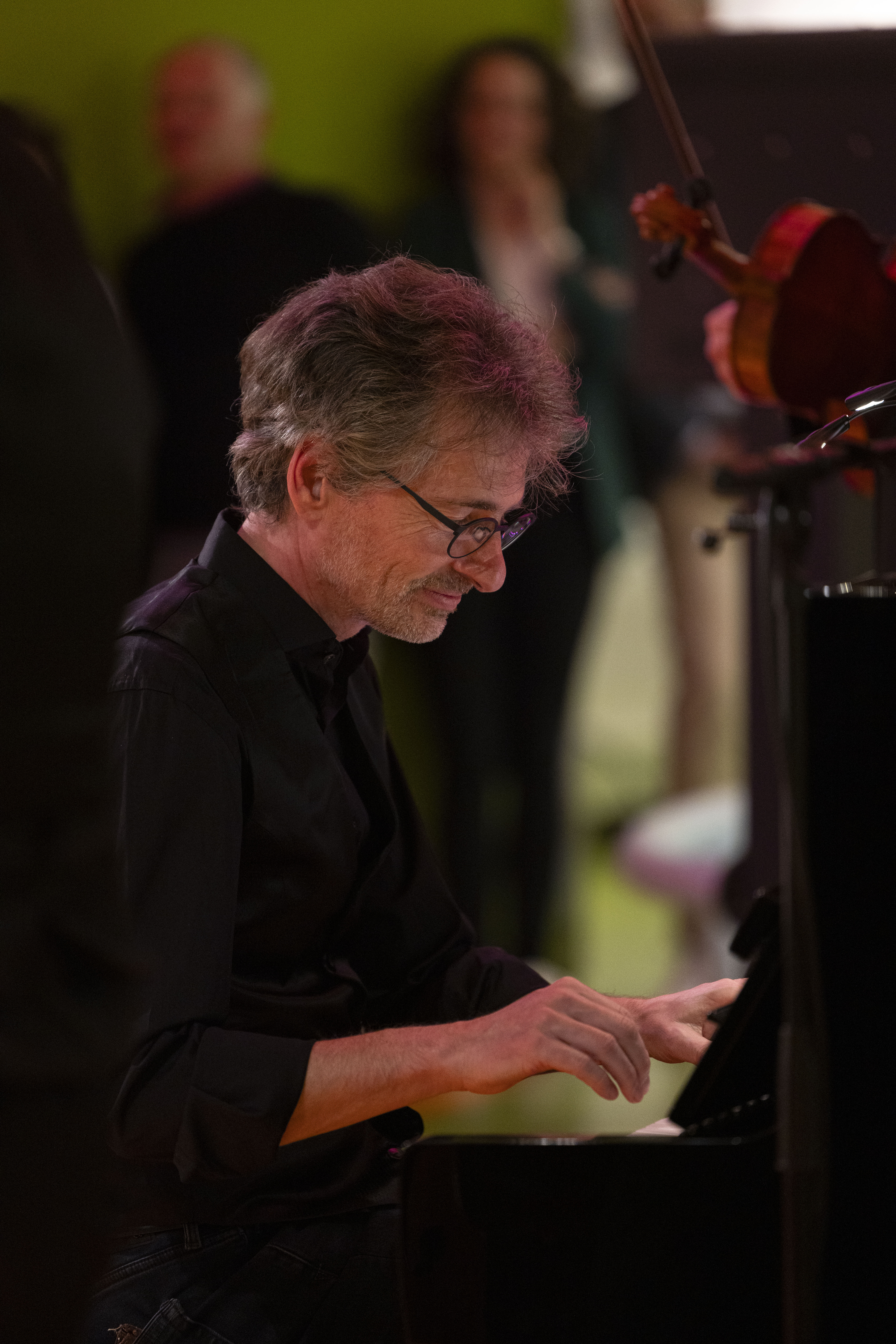 Raphael Haeger spielt konzentriert Klavier und trägt ein dunkles Hemd. Der Hintergrund ist unscharf und zeigt Menschen, die in einer warm beleuchteten Umgebung stehen und zusehen.