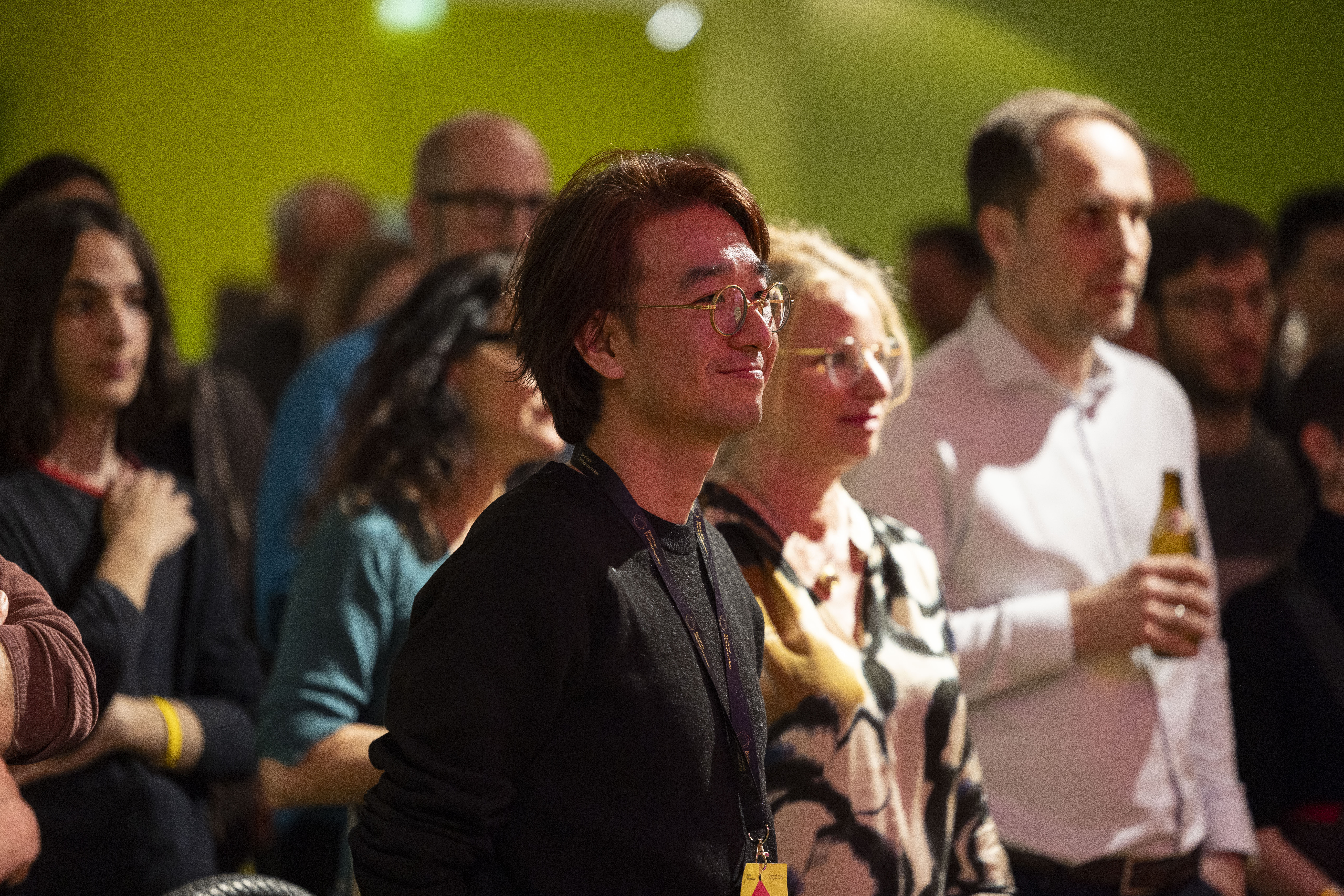 Eine Gruppe von Menschen steht bei einer Indoor-Veranstaltung dicht beieinander; eine Person vorne mit Brille und Schlüsselband lächelt, während die anderen aufmerksam zusehen. Eine Person hält eine Bierflasche in der Hand.