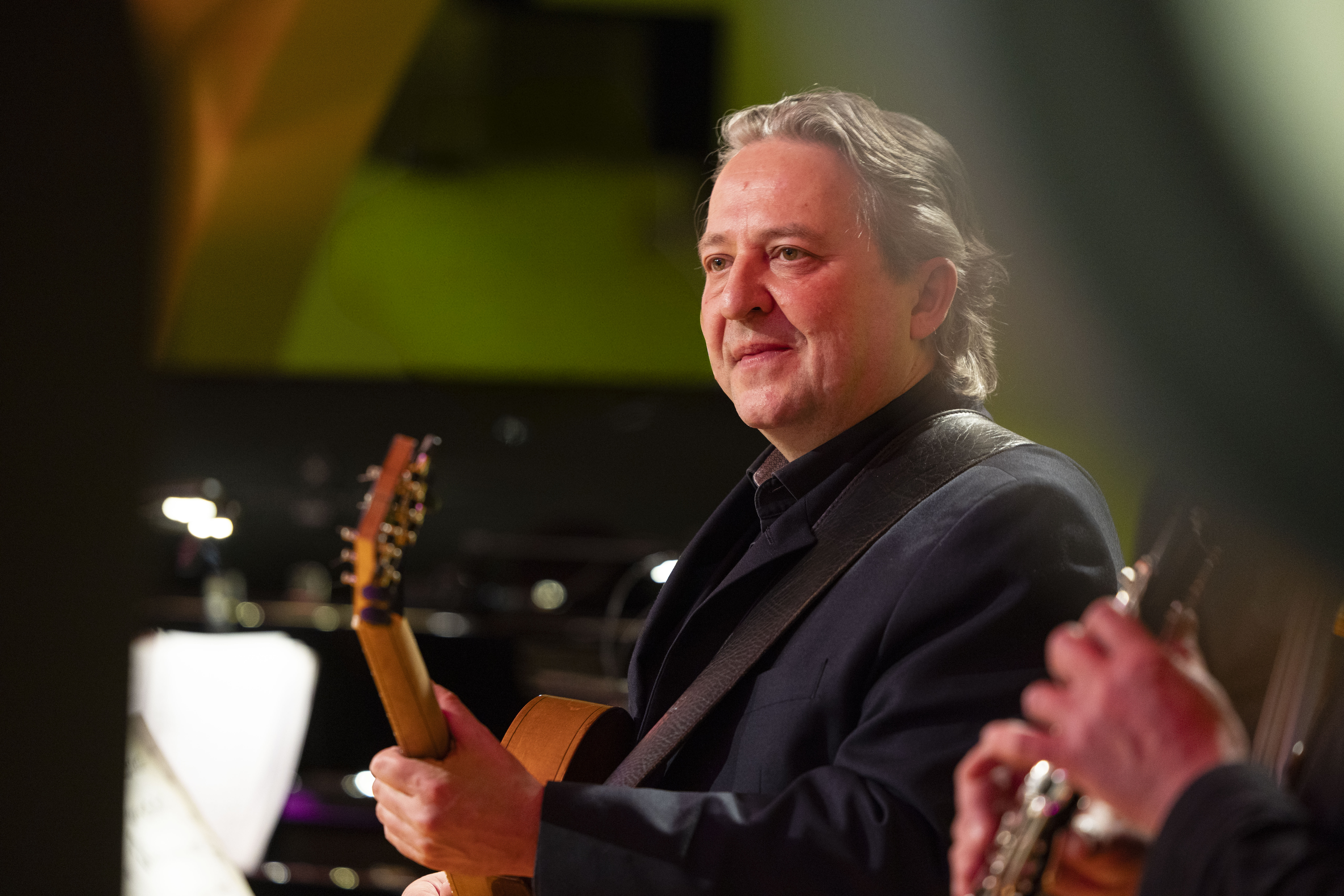 Ein Mann mittleren Alters mit grauem Haar lächelt, während er in einem dunklen Anzug auf der Bühne eine Gitarre spielt. Eine andere Person, die eine Gitarre spielt, ist teilweise im Vordergrund zu sehen.