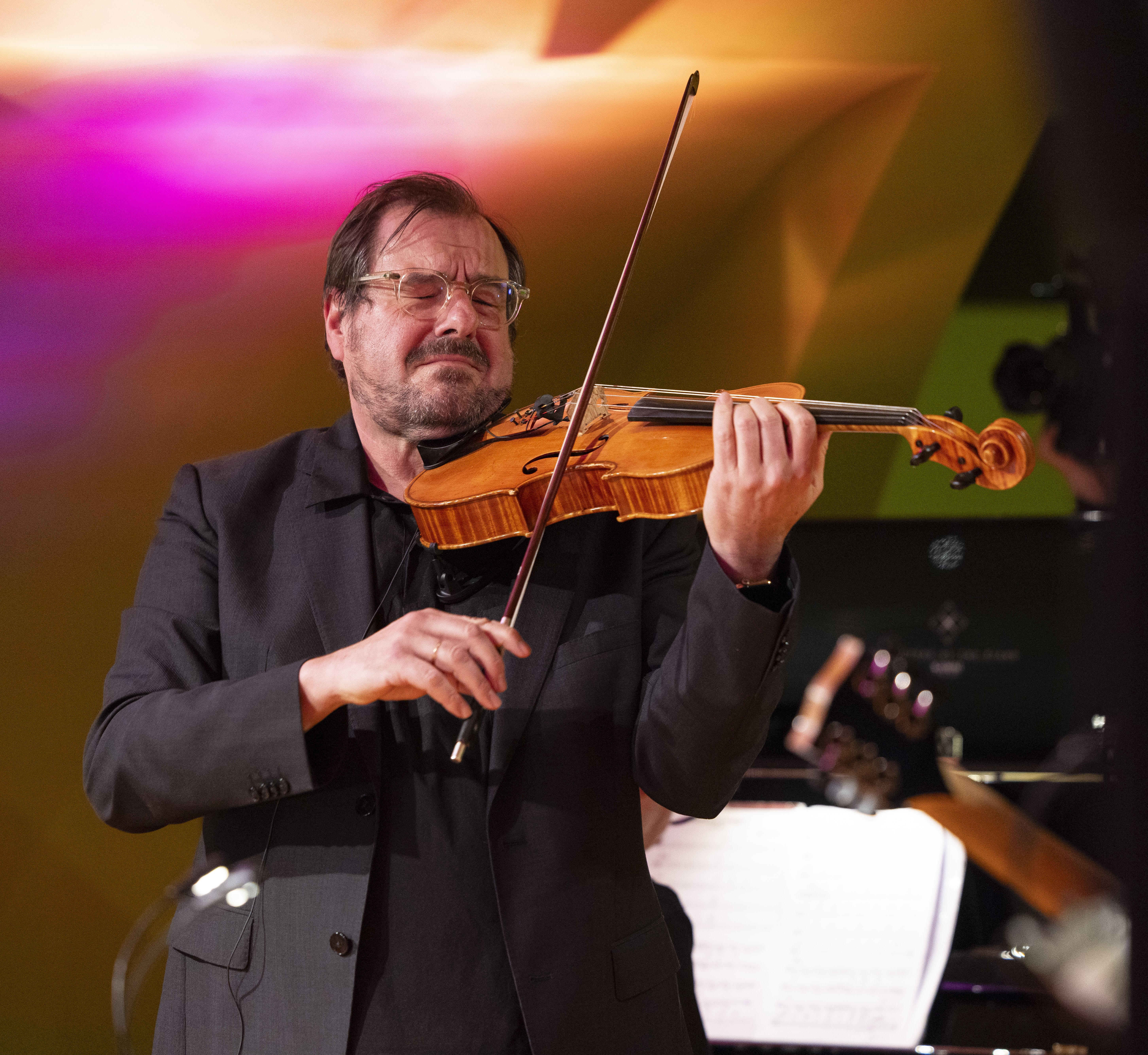 Martin Stegner, der einen dunklen Anzug trägt, spielt auf der Bühne leidenschaftlich auf einer Bratsche. Im Hintergrund sind bunte Lichter und verschwommene Musikinstrumente zu sehen.