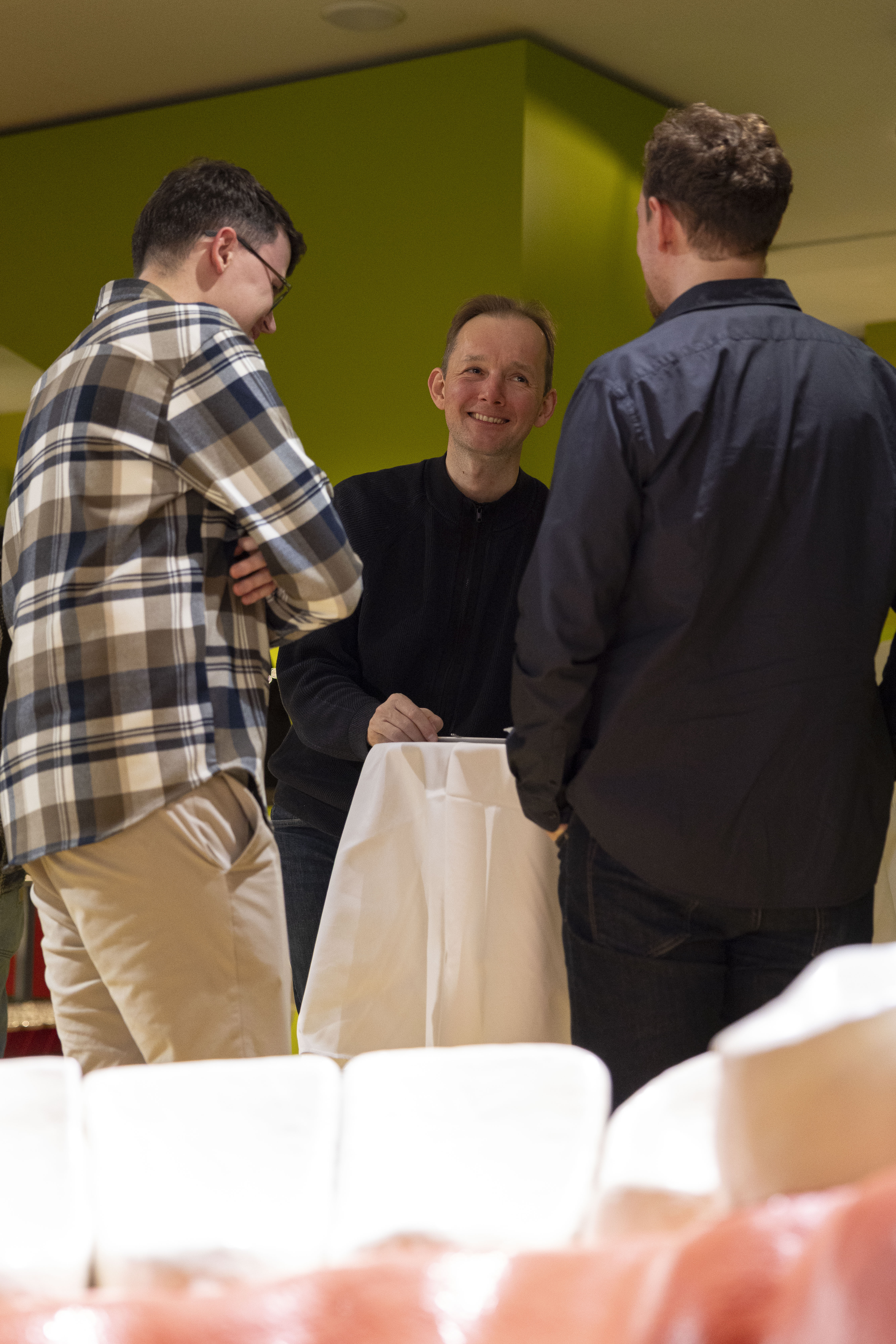 Drei Männer stehen um einen hohen runden Tisch, der mit einem weißen Tuch bedeckt ist, und unterhalten sich in einem Innenraum. Ein Mann in der Mitte lächelt. Die Wand im Hintergrund ist grün.