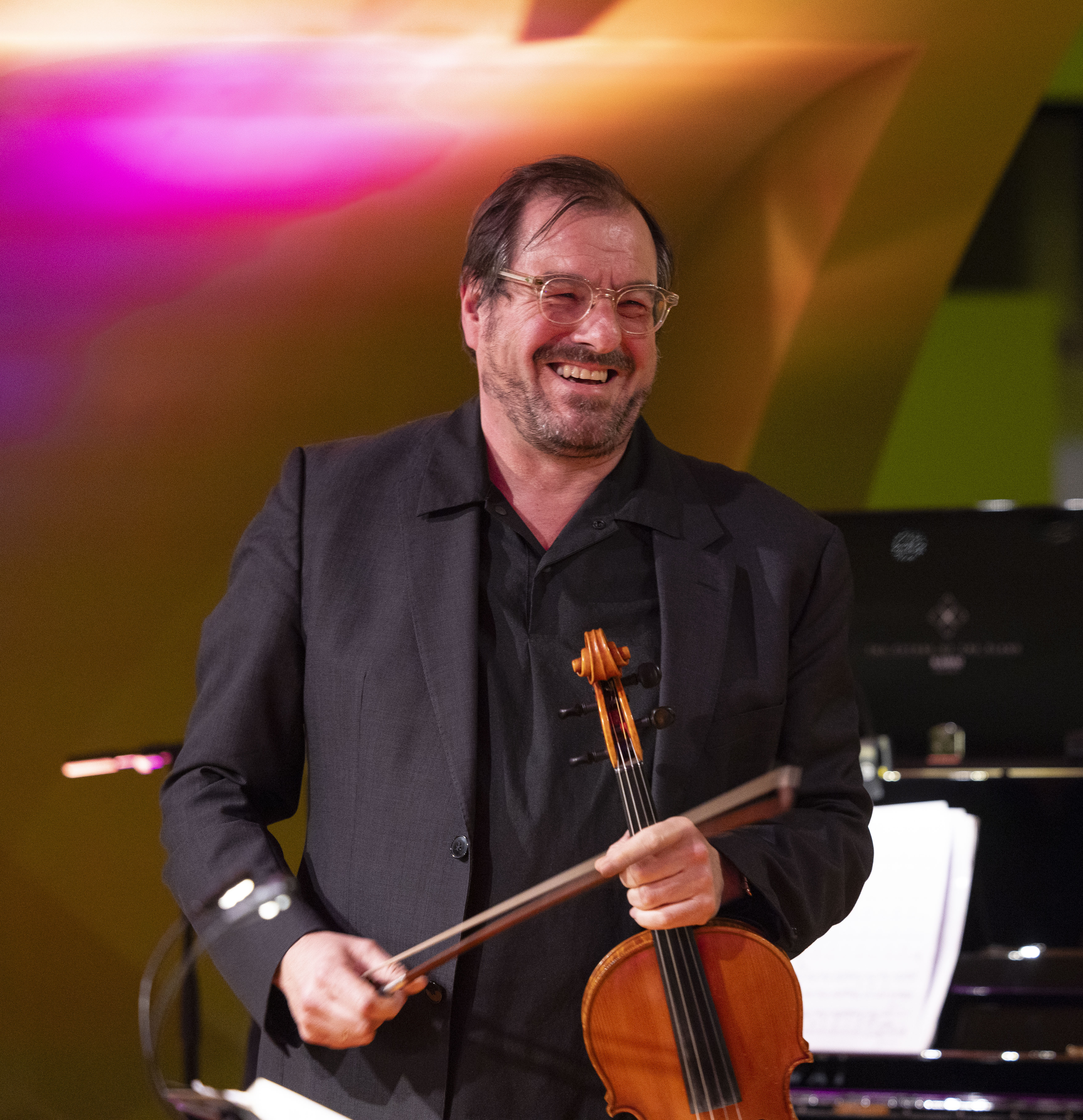 Martin Stegner in dunklem Anzug hält eine Bratsche und einen Bogen und steht vor einem Klavier mit bunten Lichtern im Hintergrund.