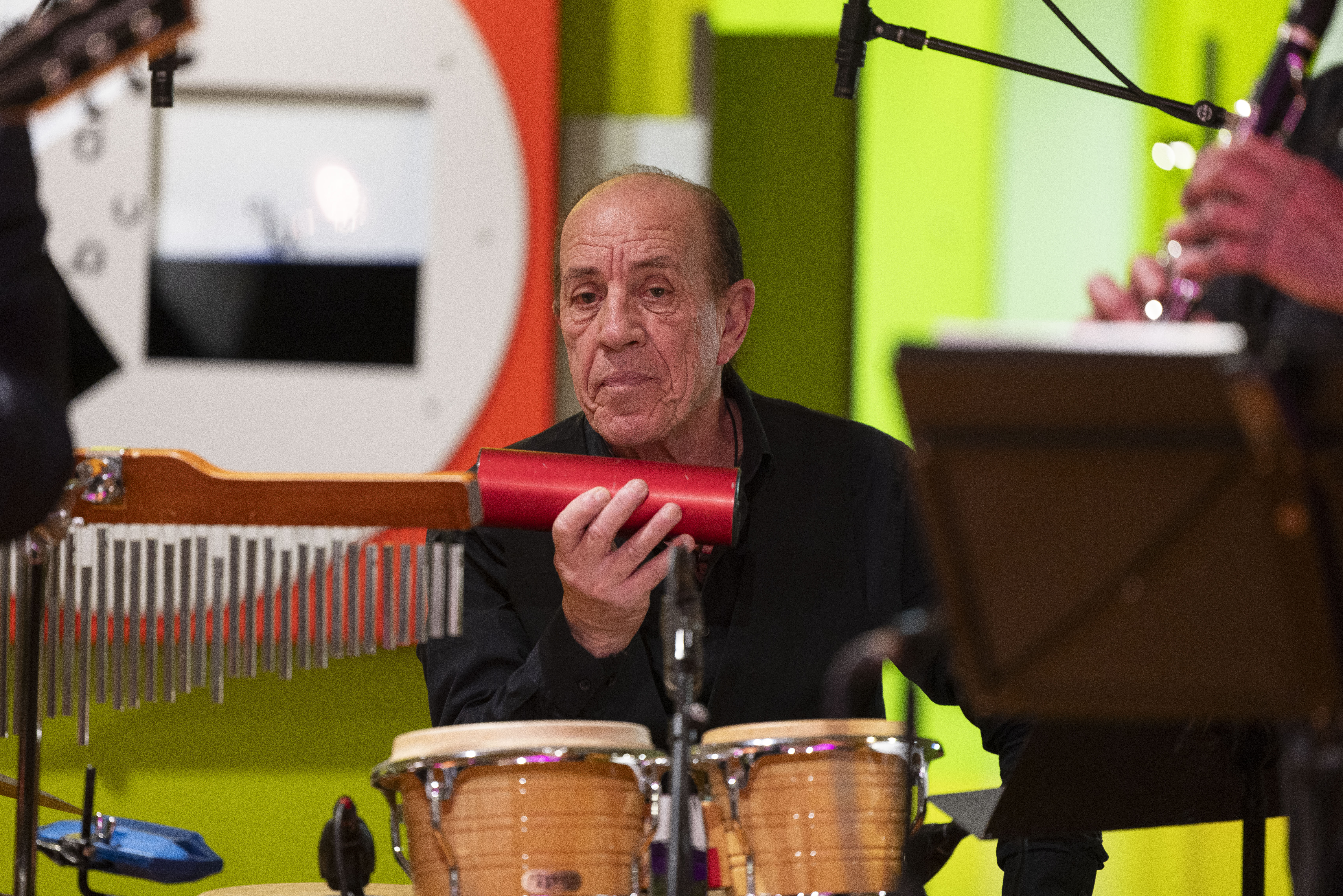 Ein älterer Mann spielt Schlaginstrumente, hält einen roten Shaker in der Hand und sitzt hinter zwei Conga-Trommeln, während um ihn herum bunte Wände und andere Musiker zu sehen sind.
