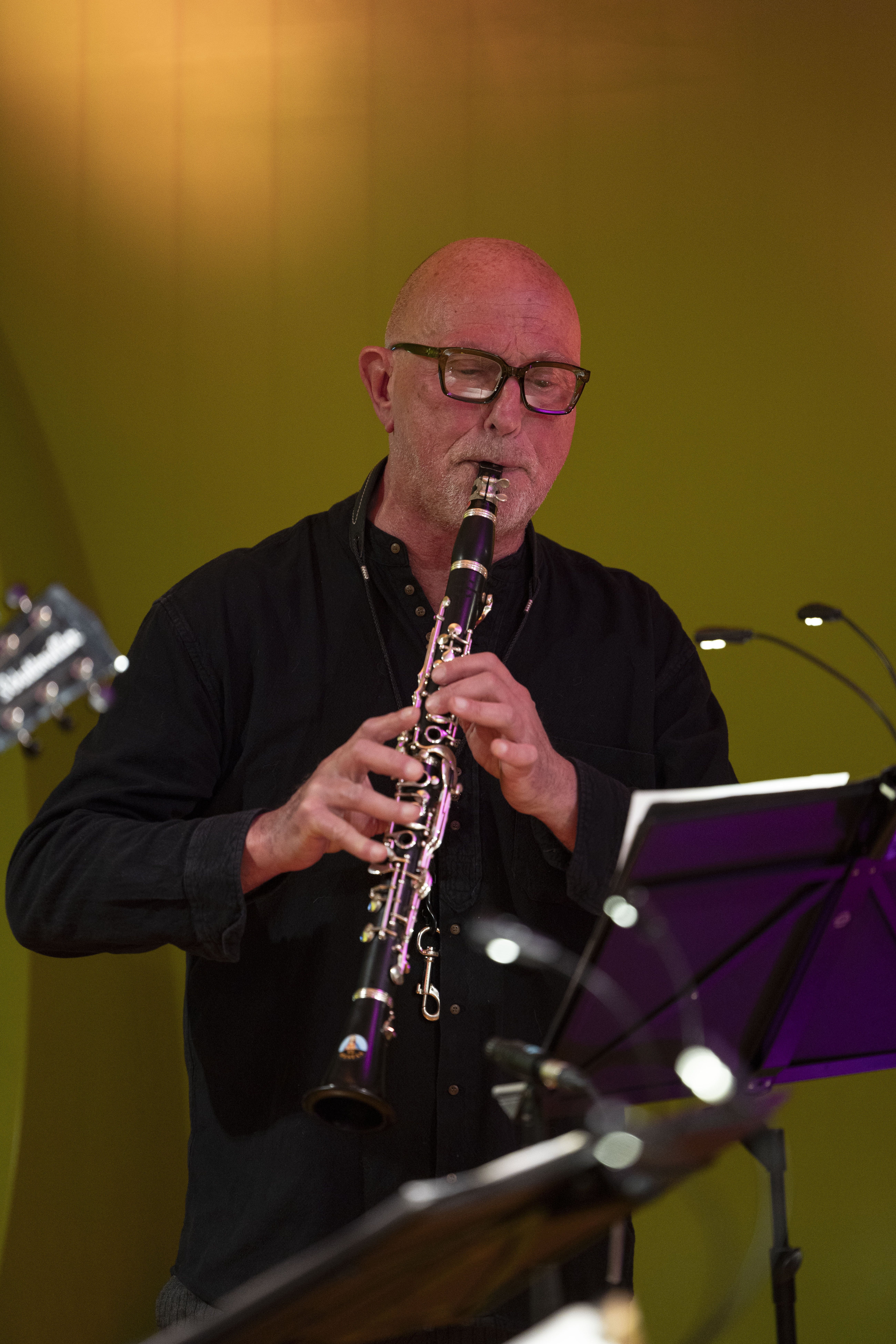 Ein Mann mit Brille spielt im Stehen Klarinette und liest Noten aus einem Ständer, vor einem schlichten gelb-grünen Hintergrund.