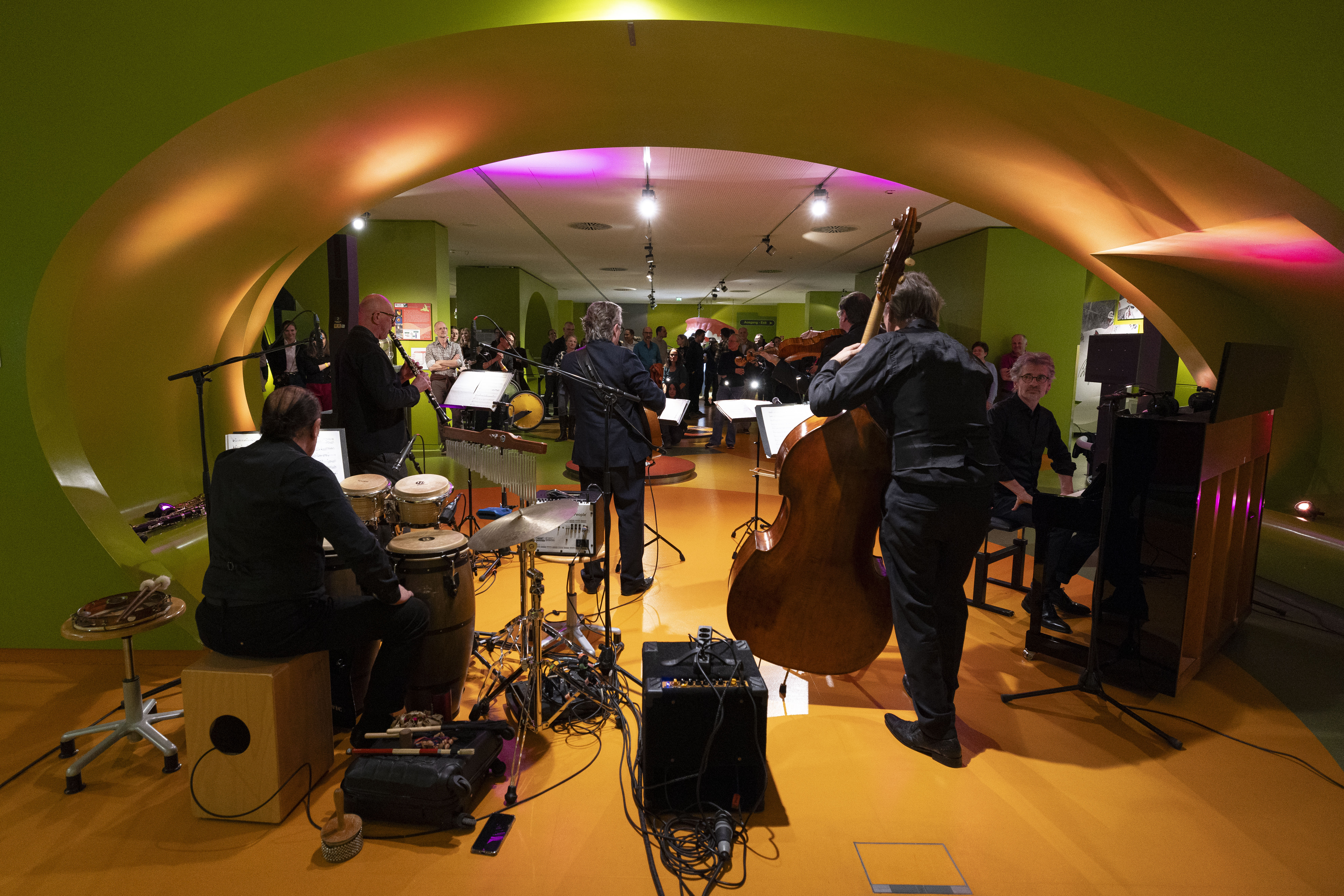 Auf der Bühne unter den geschwungenen grünen Bögen spielt eine Jazzband mit Musikern, die Schlagzeug, Kontrabass und Saxophon spielen, während das Publikum im warm beleuchteten Hintergrund zusieht.