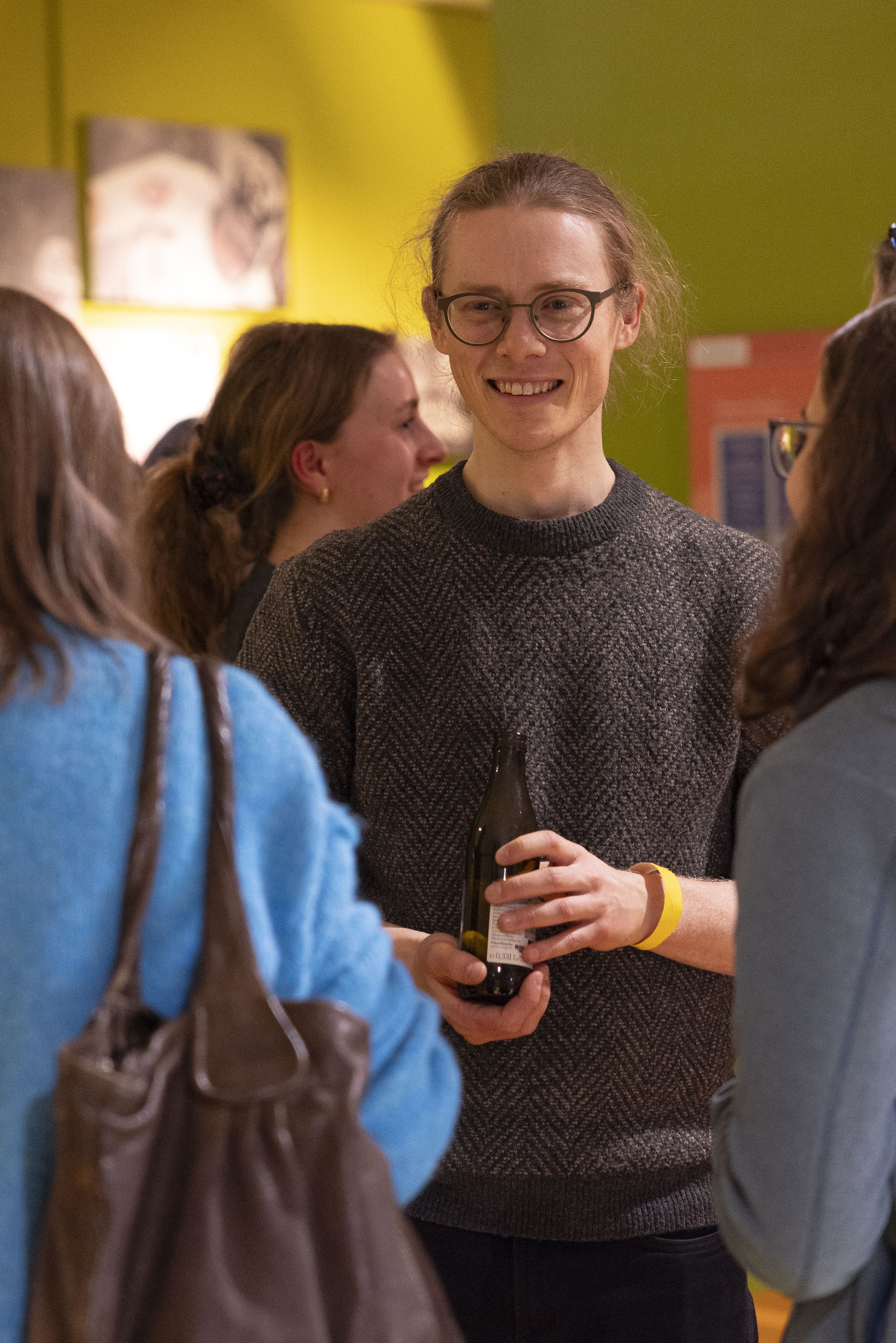 Eine lächelnde Person mit Brille und grauem Pullover hält ein Getränk in der Hand, während sie sich mit anderen bei einem geselligen Beisammensein in einem warm beleuchteten Raum unterhält.