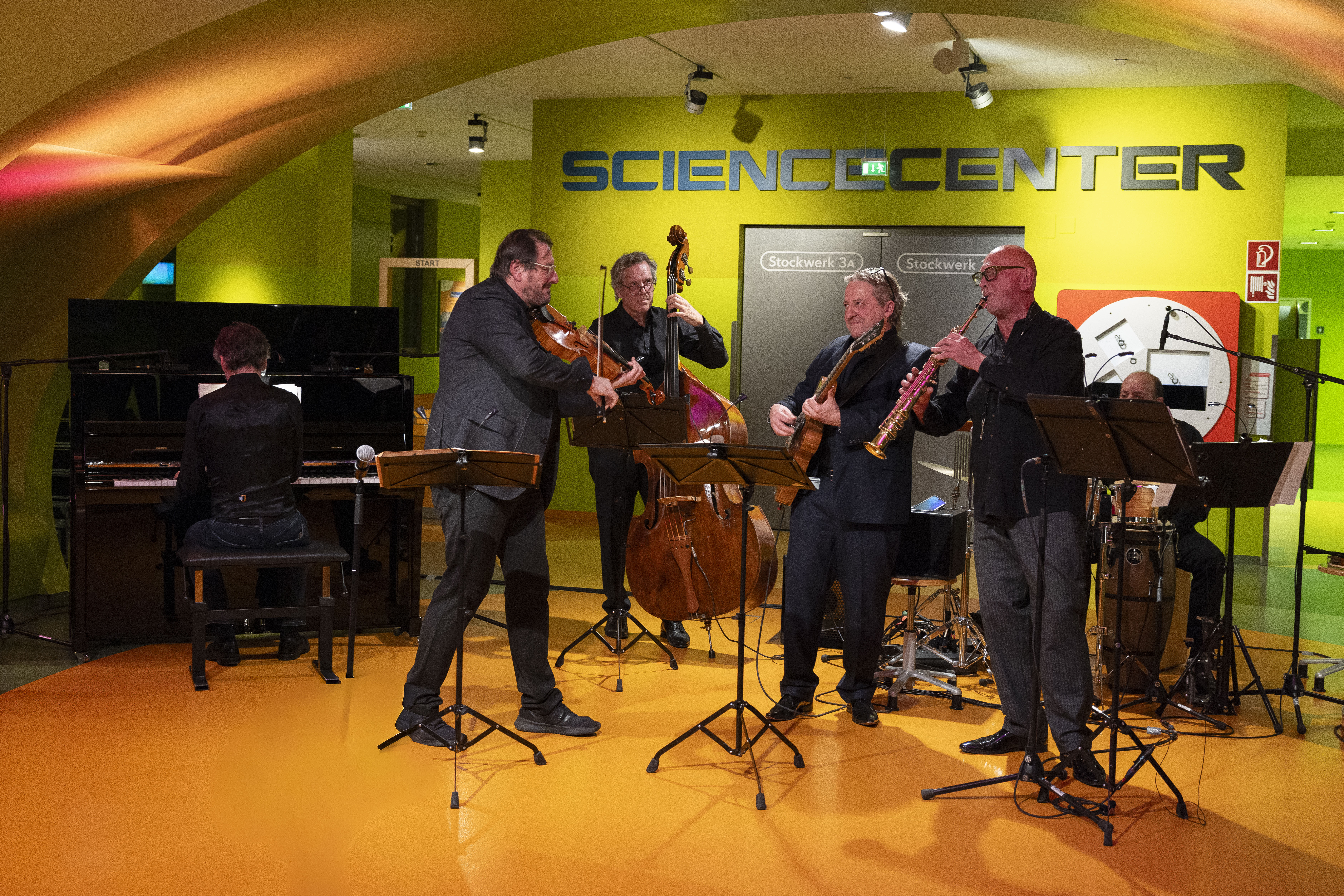Fünf Musiker spielen in einem Raum mit Klavier, Bratsche, Kontrabass, Klarinette, Gitarre und Schlagzeug vor einer grünen Wand mit der Aufschrift SCIENCE CENTER. Notenständer und Instrumente sind sichtbar.
