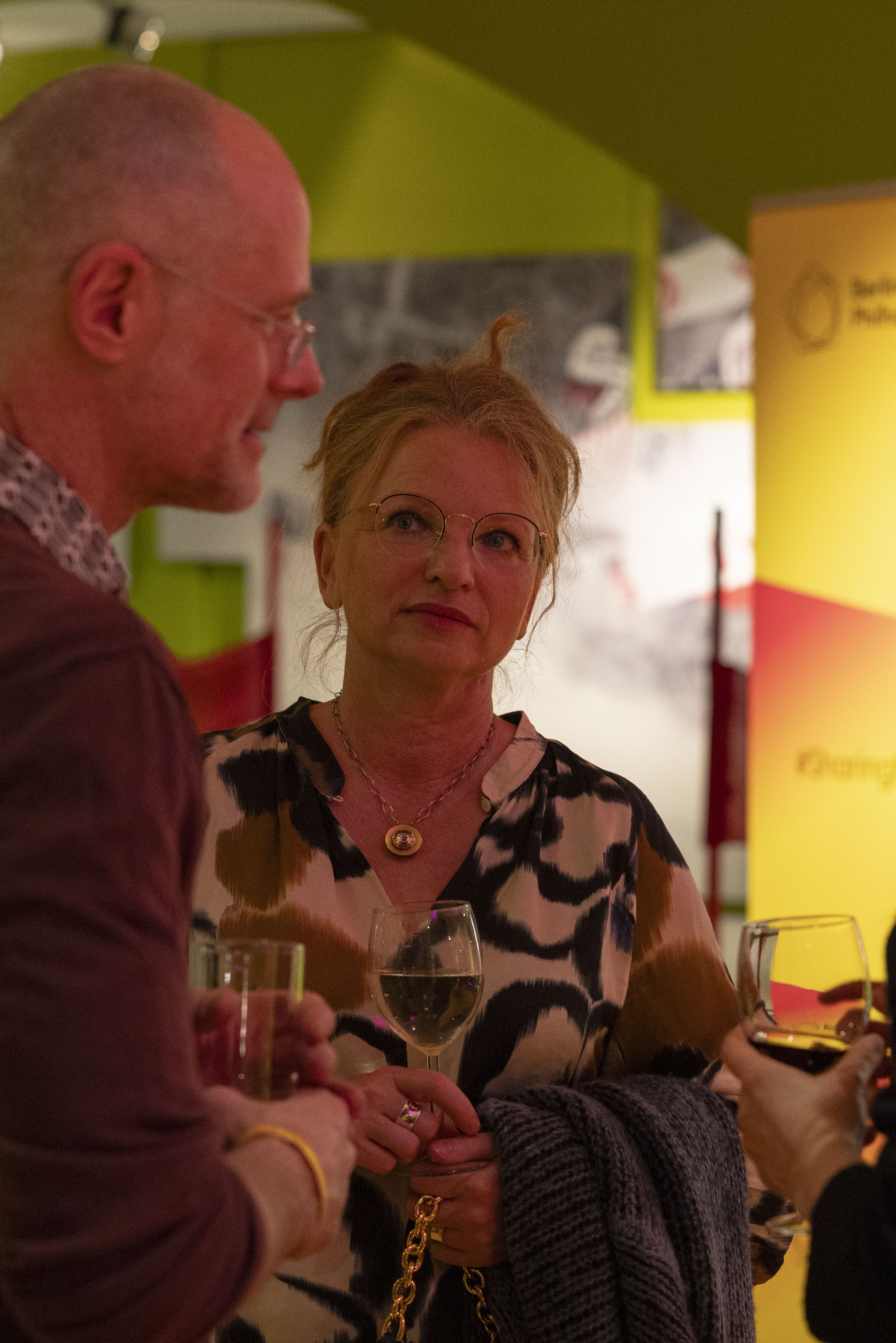 Eine Frau mit Brille und gemusterter Bluse hält ein Glas Wein in der Hand. Sie steht zwischen zwei Personen bei einer gesellschaftlichen Veranstaltung mit warmer Beleuchtung und buntem Hintergrund.