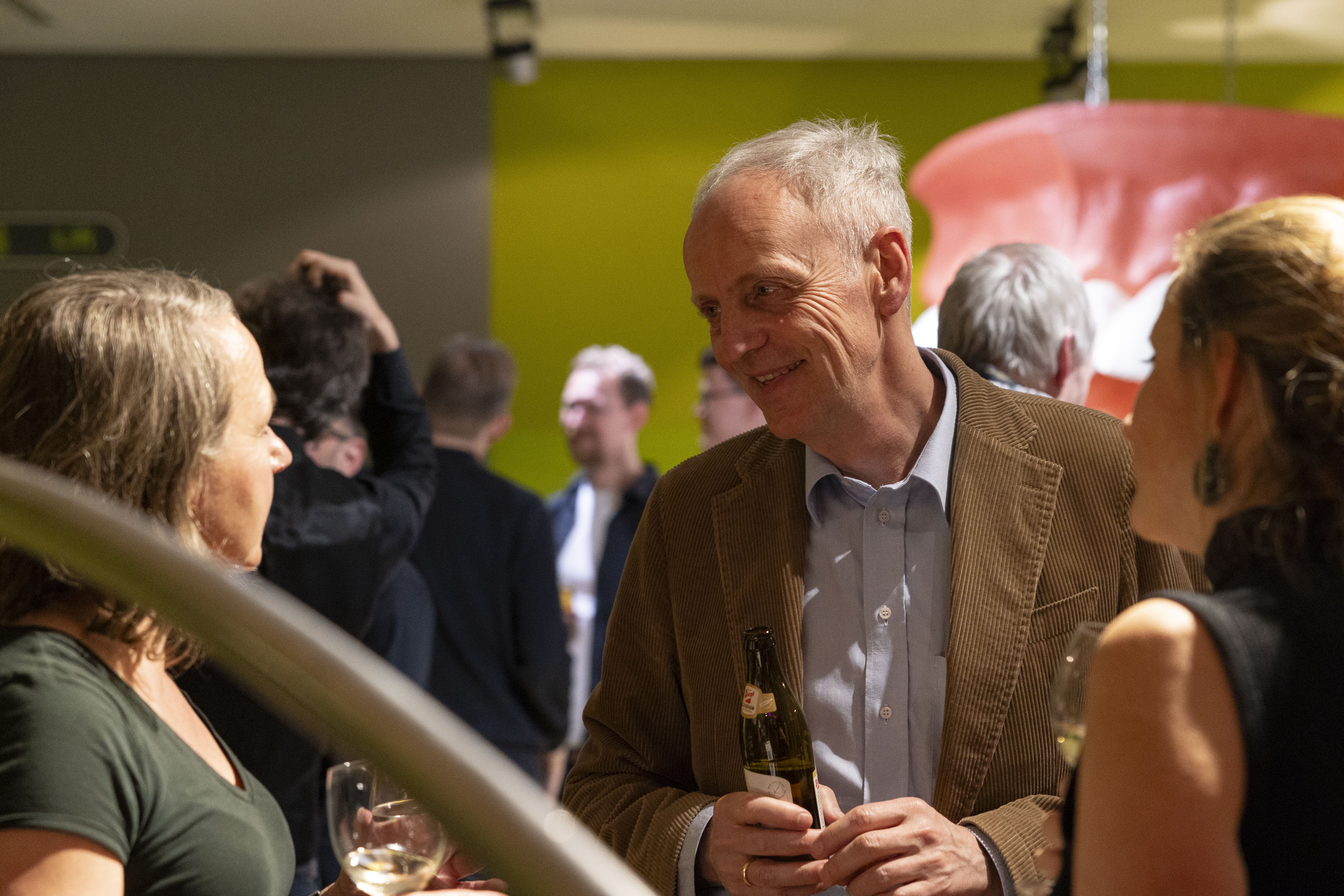 Eine Gruppe von Menschen unterhält sich bei einem gesellschaftlichen Ereignis und lächelt, während sie Getränke in der Hand halten. Der Fokus liegt auf einem Mann in einer braunen Jacke, der sich mit zwei Frauen unterhält. Der Hintergrund ist mit einer grünen Wand warm beleuchtet.