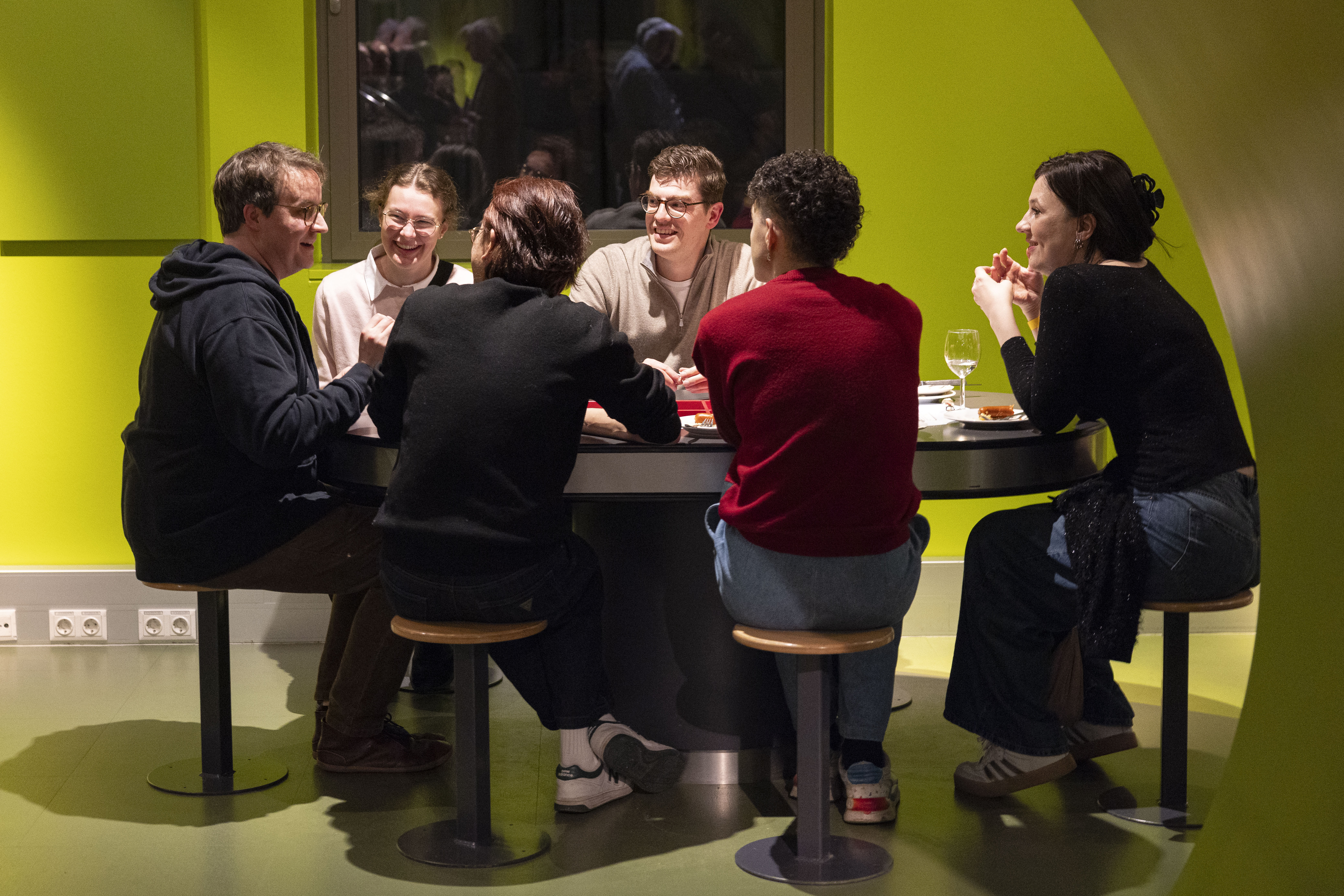 Sechs Personen sitzen in einem hell erleuchteten Raum mit gelb-grünen Wänden um einen runden Tisch, unterhalten sich und lächeln. Auf dem Tisch stehen Speisen und Getränke, und die Gruppe scheint ein freundliches Gespräch zu führen.