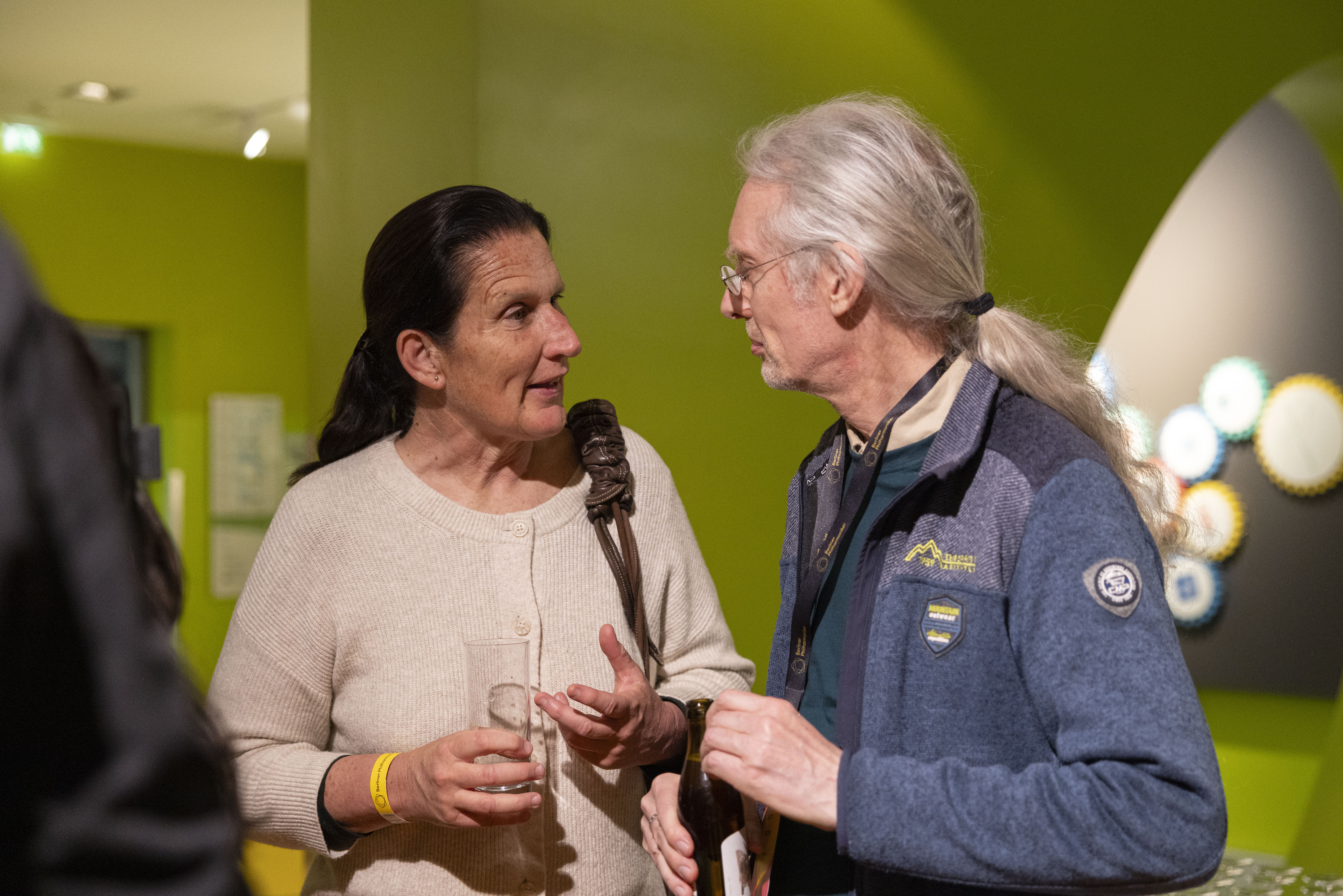 Eine Frau und ein älterer Mann mit langen grauen Haaren unterhalten sich bei einem gesellschaftlichen Ereignis. Sie hält ein Glas in der Hand und trägt einen cremefarbenen Pullover; er trägt eine Brille und eine blaue Jacke. Der Hintergrund ist grün.