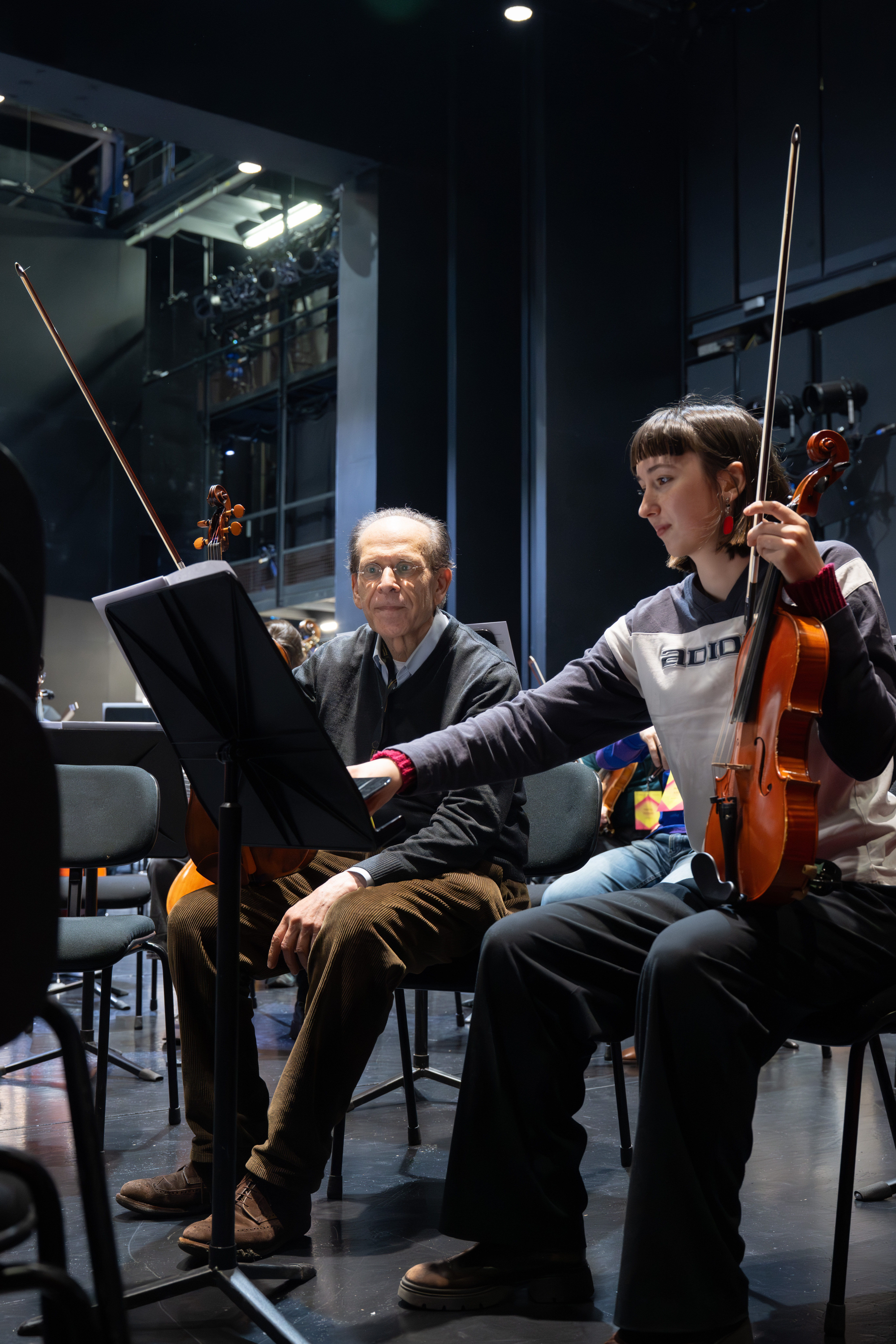 Ein älterer Mann und eine junge Frau sitzen mit Streichinstrumenten auf der Bühne. Die Frau gestikuliert auf Notenblättern, während der Mann zusieht. Beide wirken konzentriert, hinter ihnen sind Bühnenbeleuchtung und Ausrüstung zu sehen.