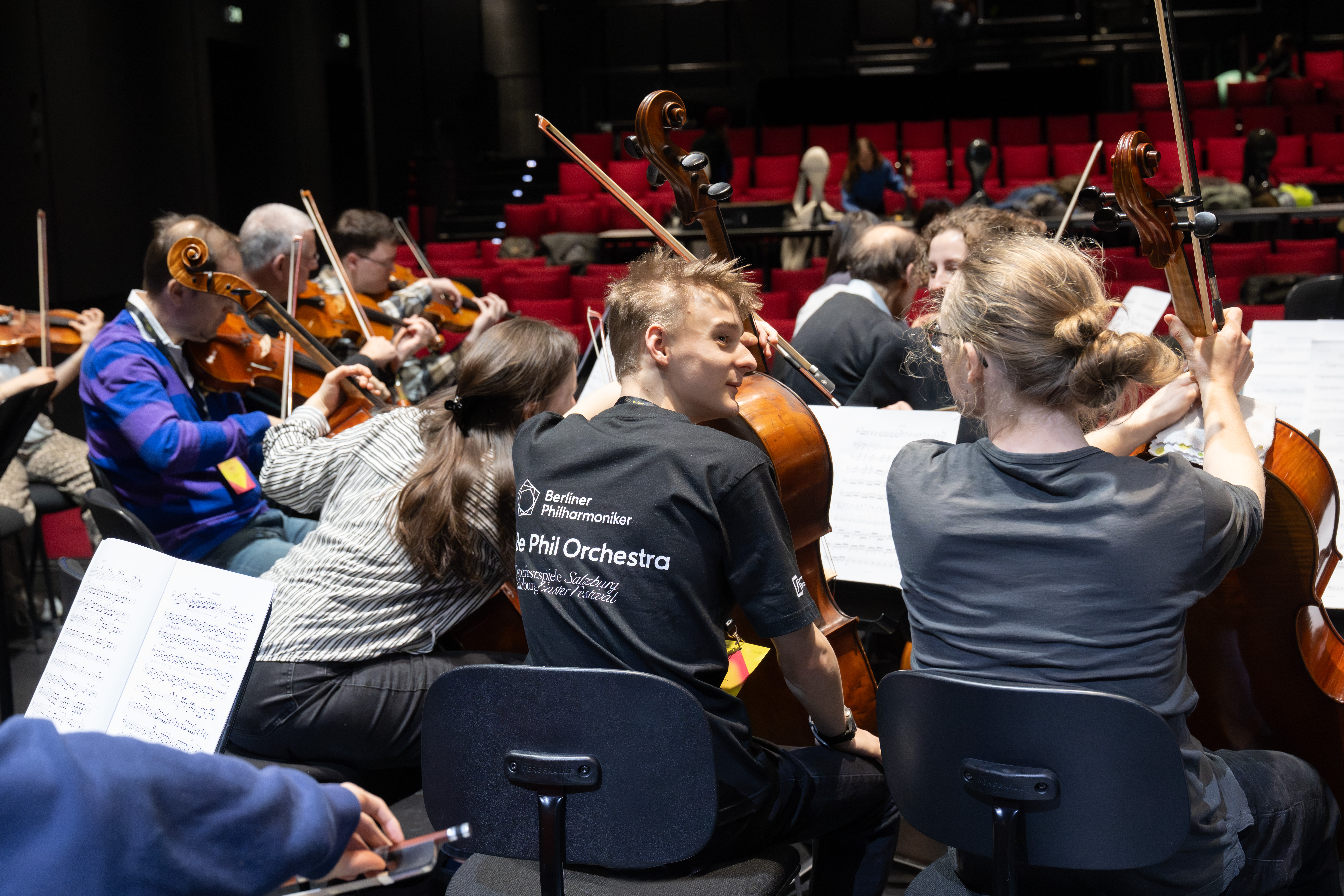 Musiker*innen eines Orchesters proben mit Streichinstrumenten auf der Bühne, in Reihen sitzend mit Notenblättern auf Ständern. Der Saal ist mit roten Sitzen und einigen Menschen im Hintergrund ausgestattet.