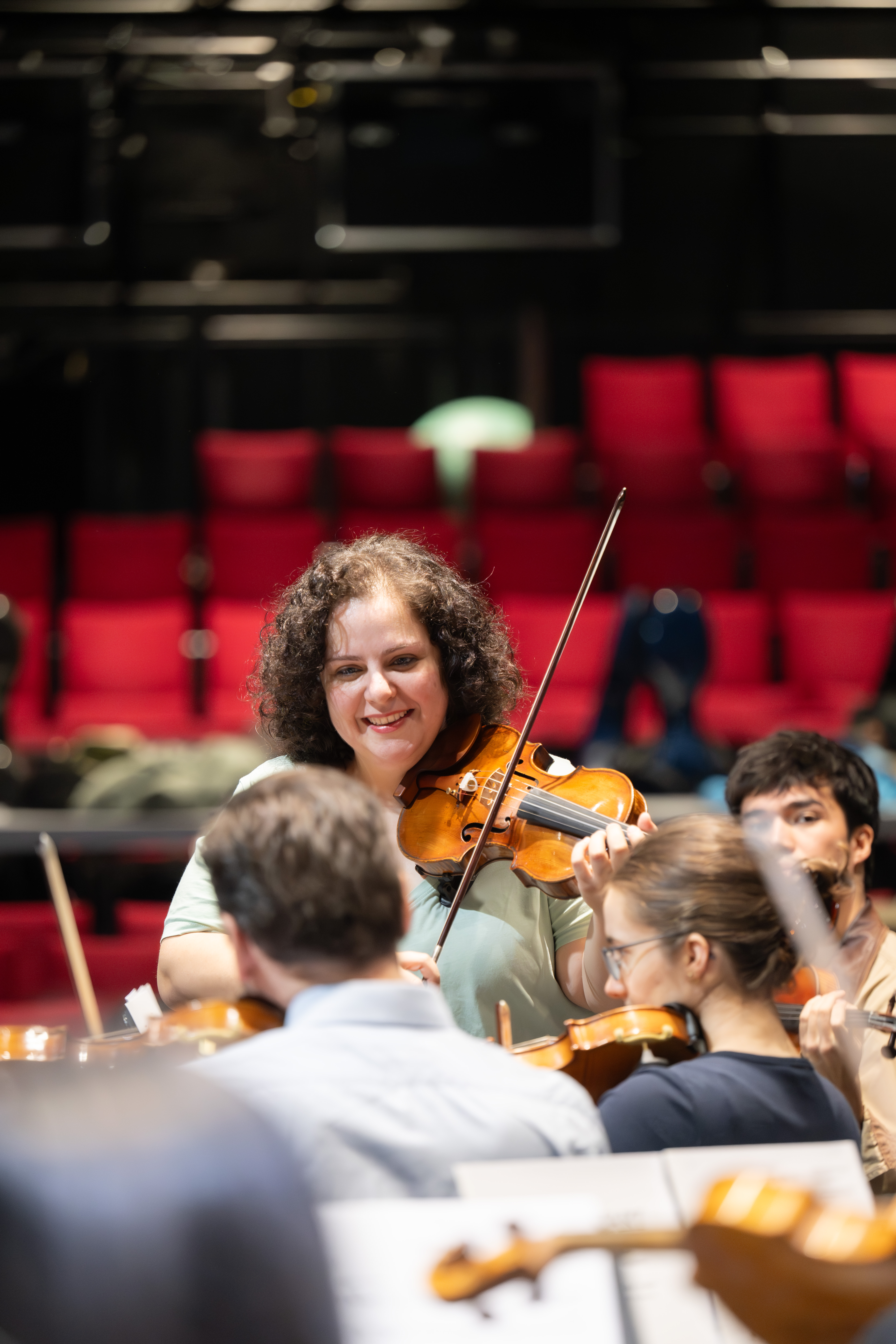 Hande Küden spielt lächelnd während einer Orchesterprobe Geige, umgeben von anderen Musikern mit Streichinstrumenten. Im Hintergrund sind rote Theatersitze zu sehen.