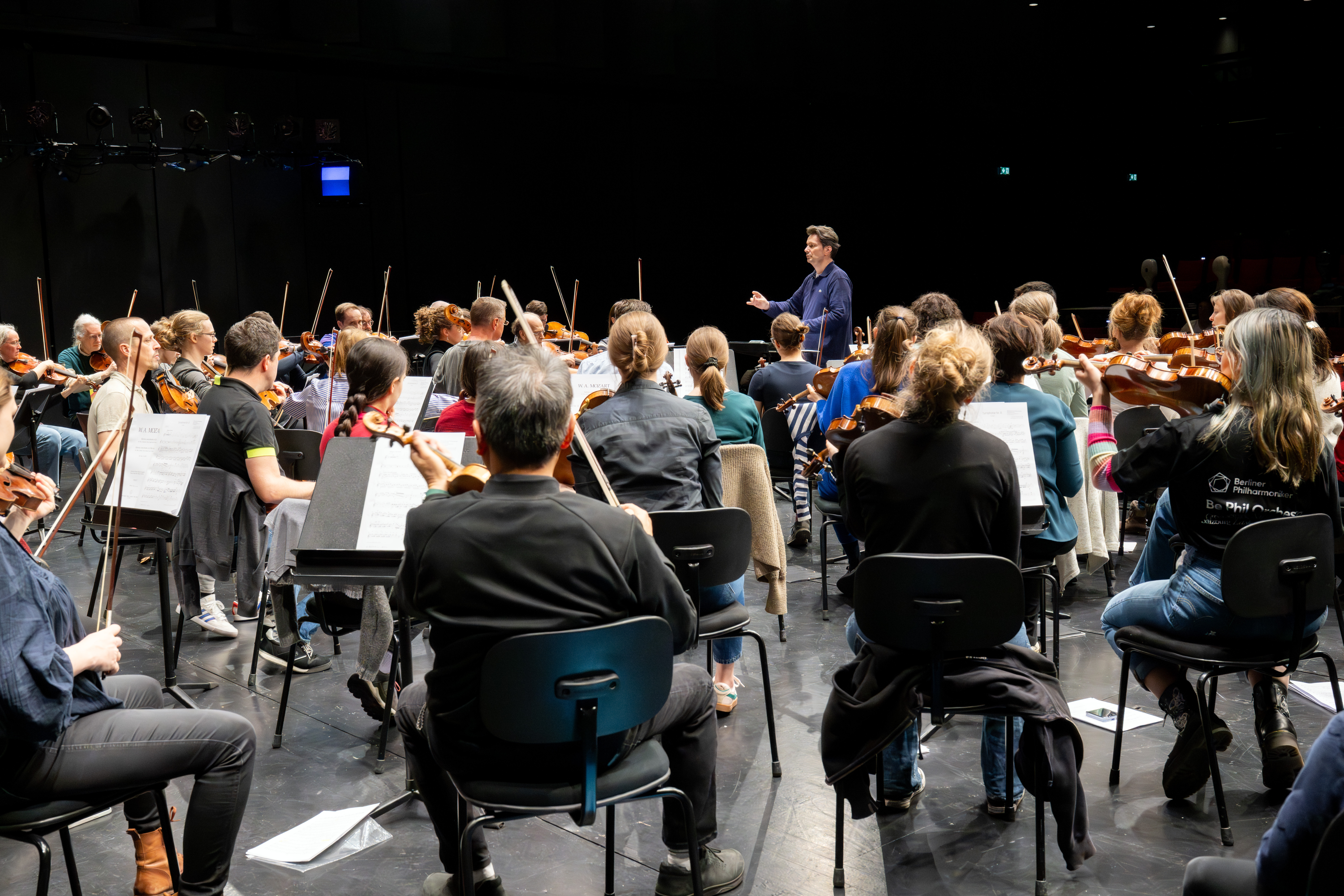 Ein Dirigent leitet eine Orchesterprobe auf der Bühne. In Reihen sitzende Musiker*innen spielen auf verschiedenen Streichinstrumenten, vor ihnen stehen Notenständer, von hinten gesehen das Orchester.