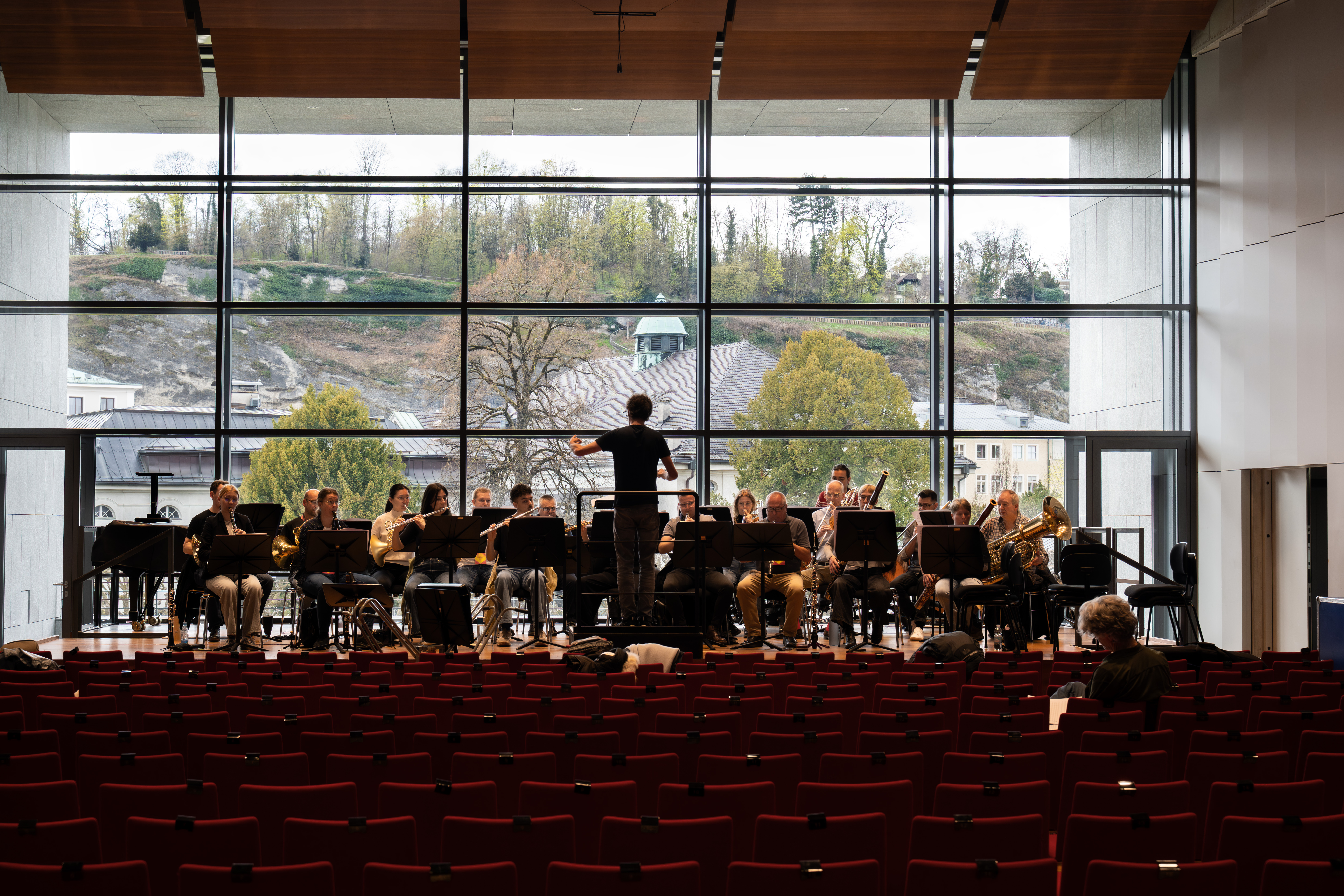 Ein Dirigent leitet eine große Orchesterprobe auf der Bühne eines modernen Konzertsaals mit raumhohen Fenstern, roten Sitzen und Bäumen im Freien. Im Publikum sitzen nur wenige Menschen.