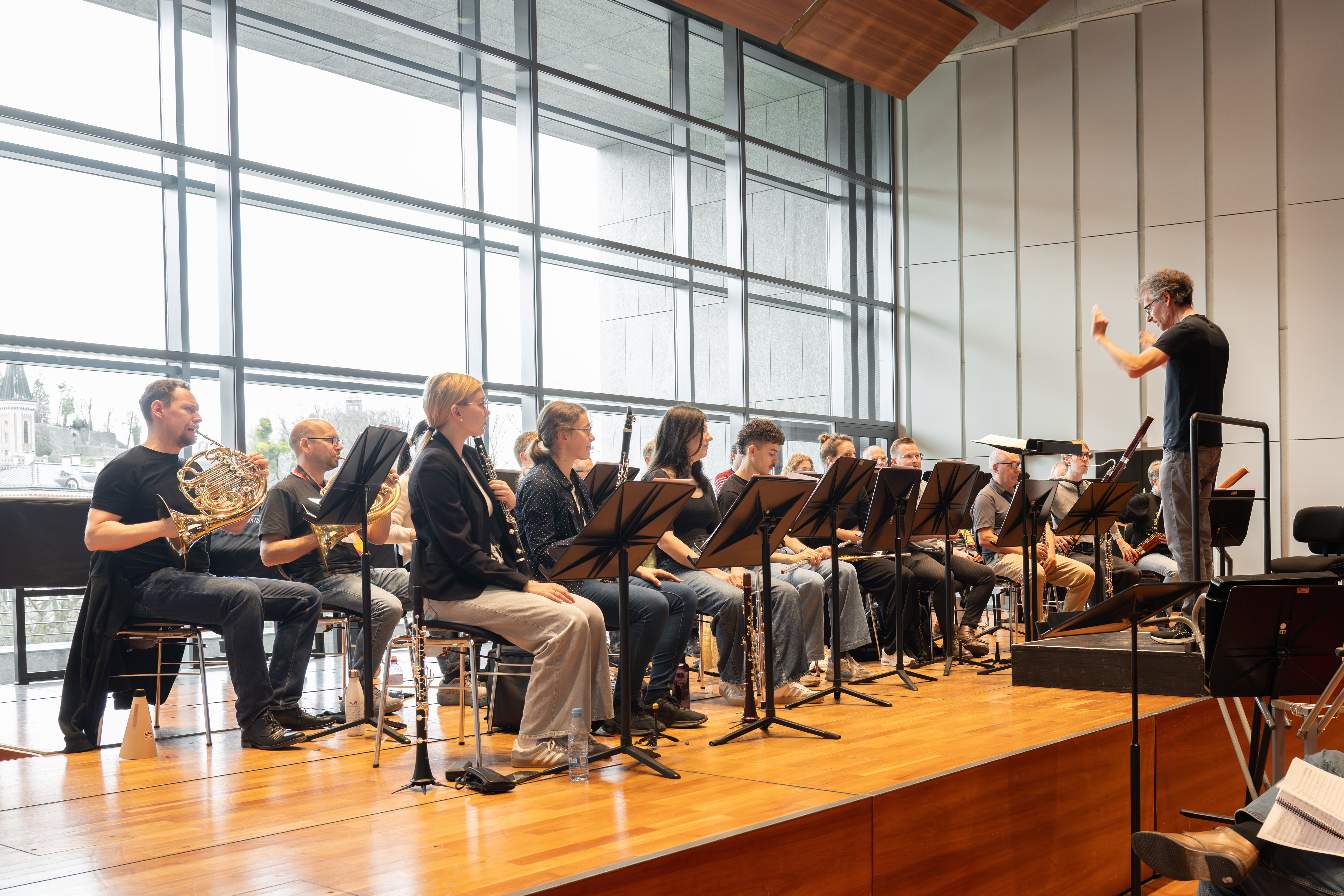 Raphael Haeger leitet eine sitzende Orchesterprobe in einer modernen, lichtdurchfluteten Halle. Die Musiker spielen verschiedene Blech- und Holzblasinstrumente und lesen von Notenständern in der Nähe großer Fenster ab.