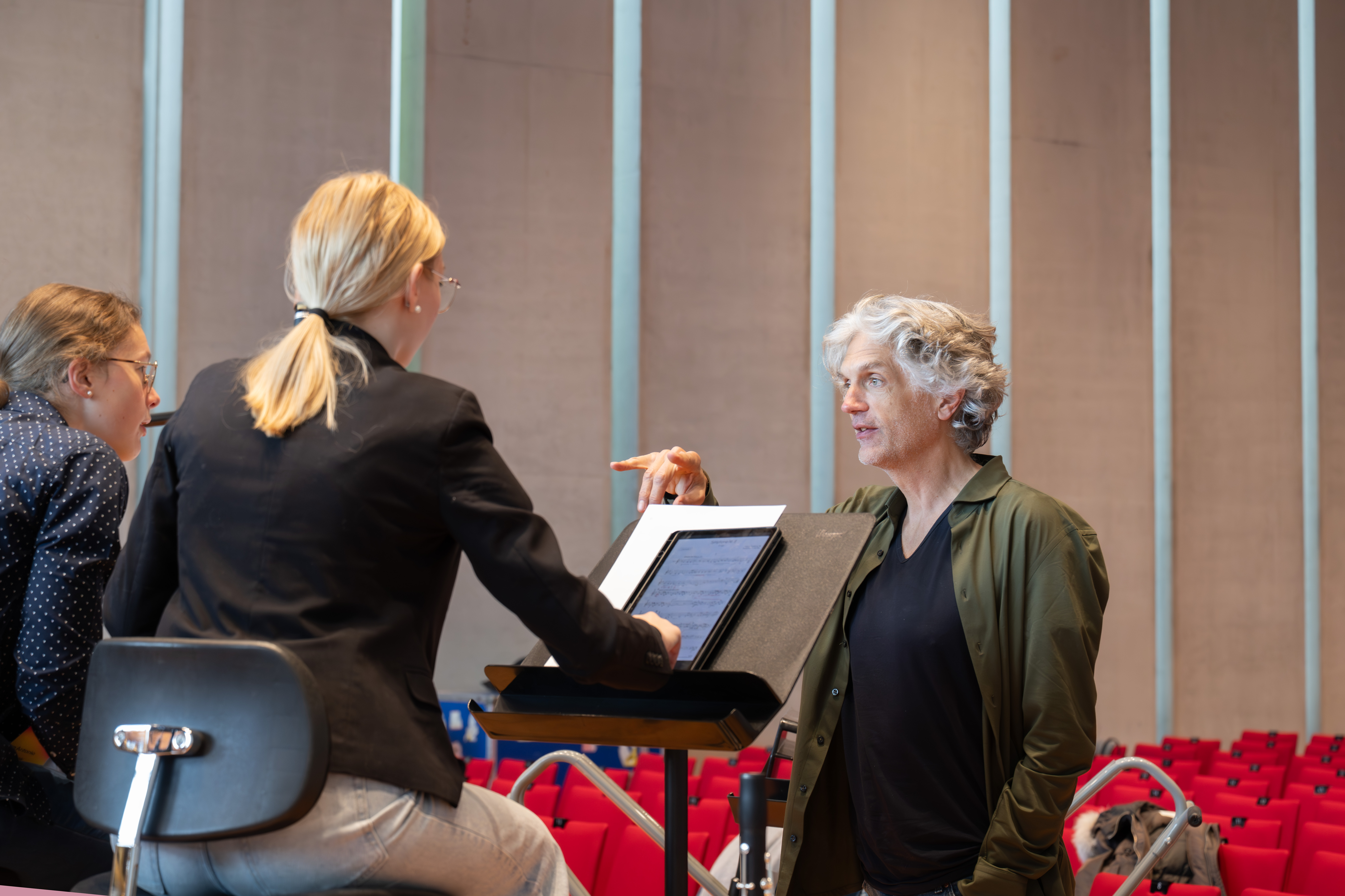 Dominik Wollenweber steht an einem Notenpult und diskutiert mit einer Musikerin in einem modernen Hörsaal mit roten Sitzen und hohen vertikalen Fenstern. Eine weitere Person sitzt in der Nähe und hört zu.