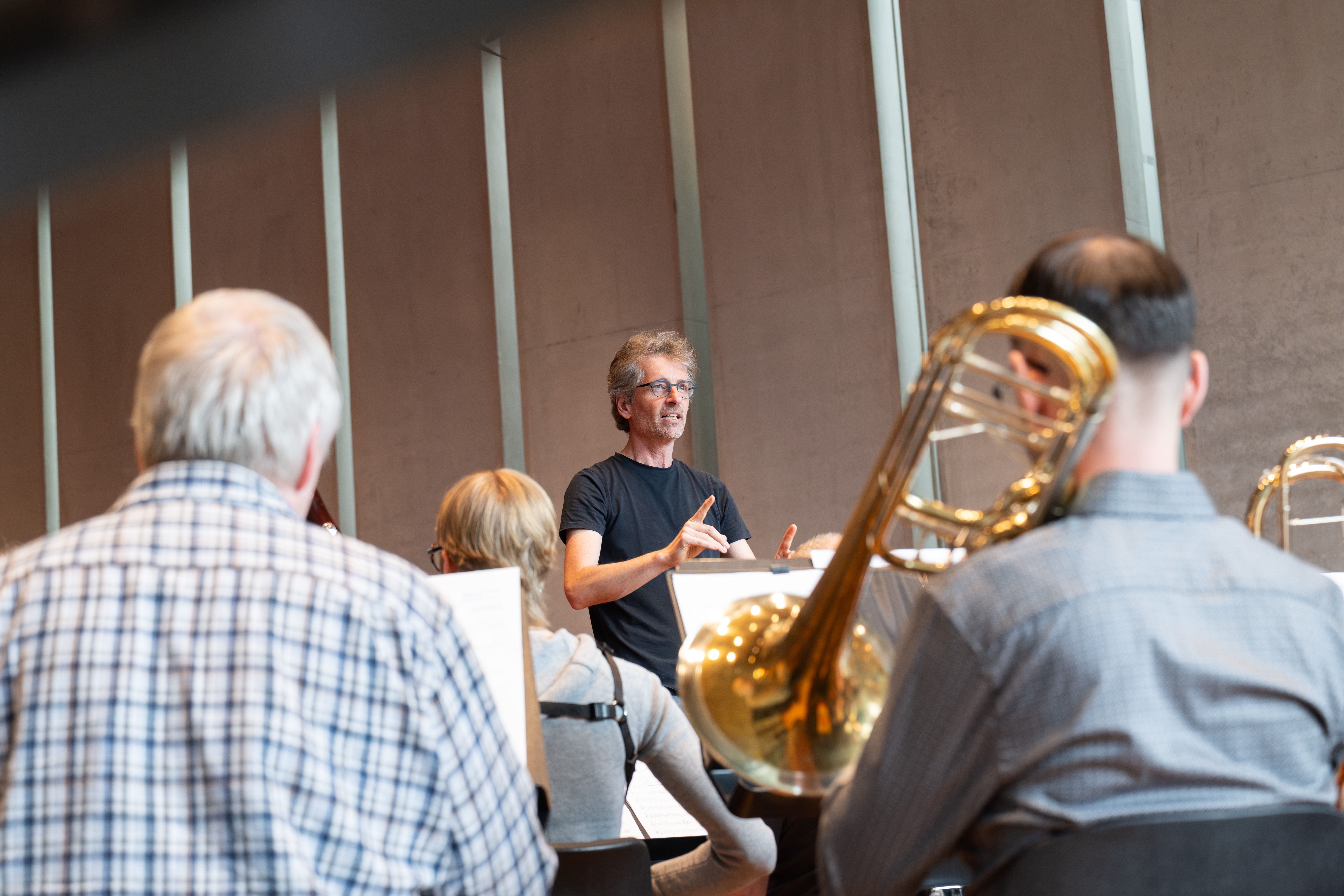 Raphael Haeger in einem schwarzen Hemd leitet eine Orchesterprobe. Die Musiker, einige mit Blechblasinstrumenten, sitzen in einem modernen, gut beleuchteten Raum mit hohen Fenstern und sind dem Dirigenten zugewandt.