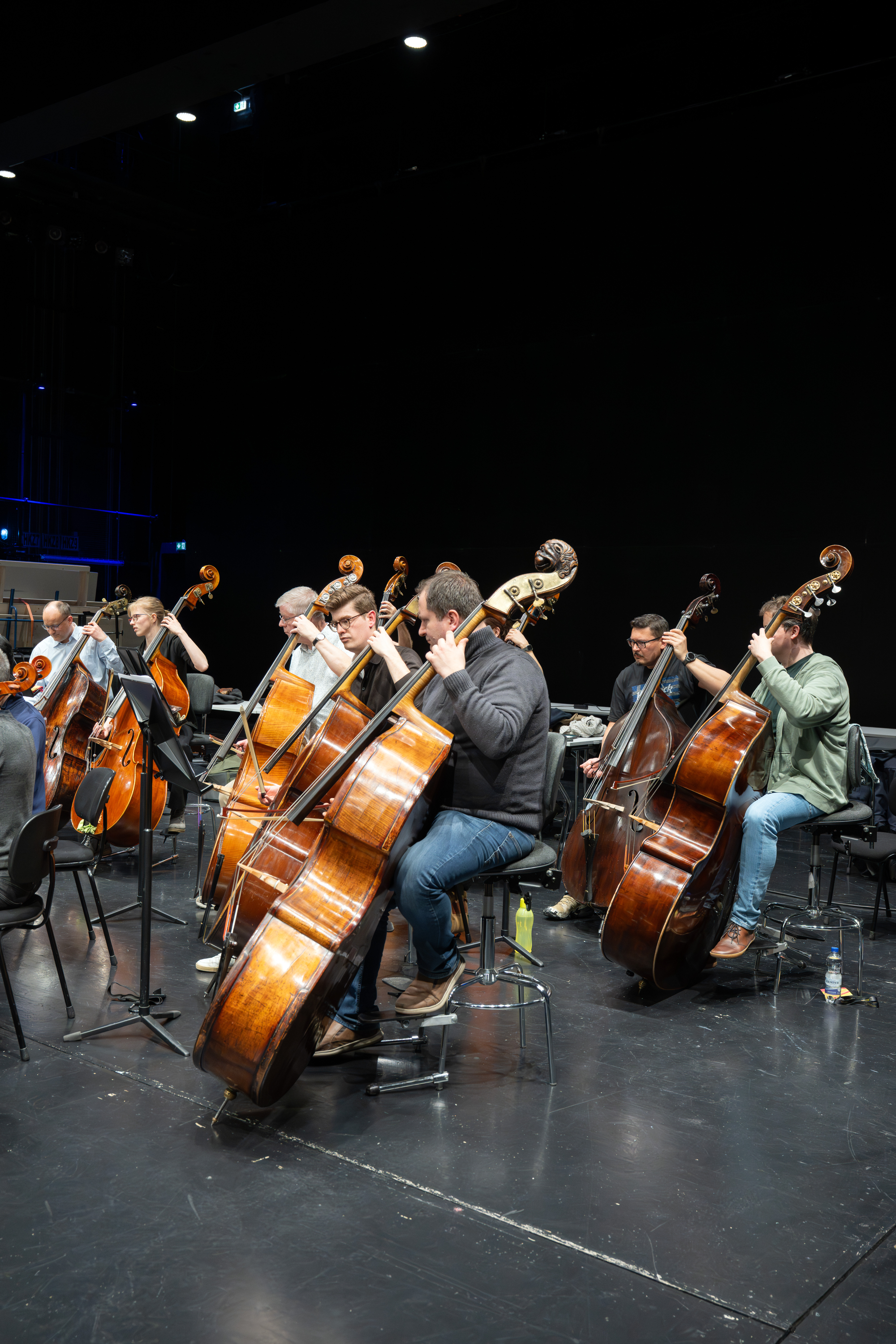 Musiker, die auf der Bühne sitzen, spielen Kontrabässe in einer Orchesterprobe. Sie konzentrieren sich auf ihre Notenblätter, und der Bühnenhintergrund ist dunkel, um die Musiker und ihre Instrumente hervorzuheben.