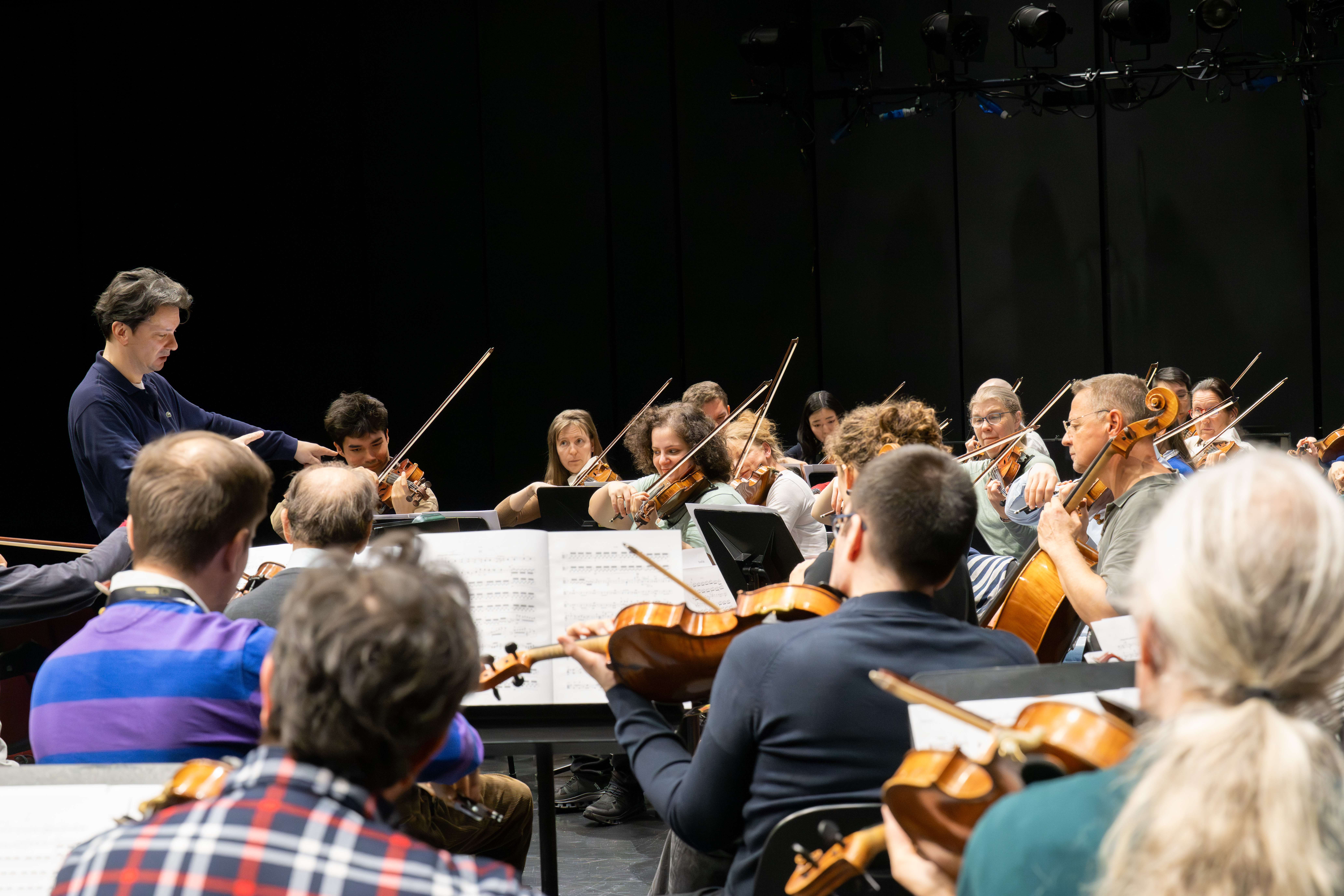 Ein Dirigent leitet ein großes Orchester während einer Probe. Die Musiker spielen Geigen, Celli und andere Streichinstrumente, während sie in einem gut beleuchteten Raum mit dunklem Hintergrund die Noten lesen.