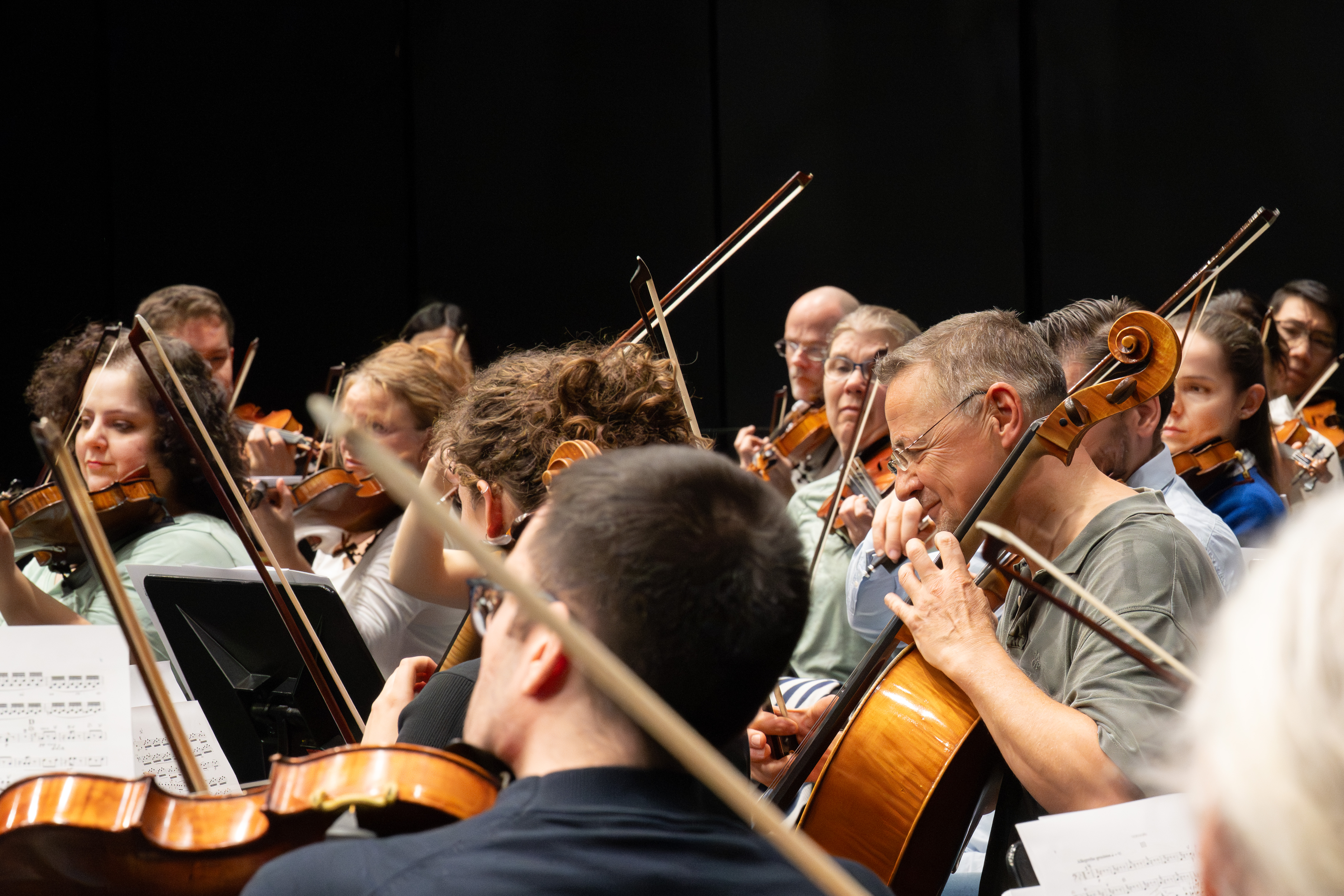 Eine Gruppe von Musiker*innen spielt Streichinstrumente, darunter Geigen und Celli, während einer Orchesterprobe. Die Noten sind zu sehen, und der Fokus liegt auf den intensiven Gesichtsausdrücken der Musiker.